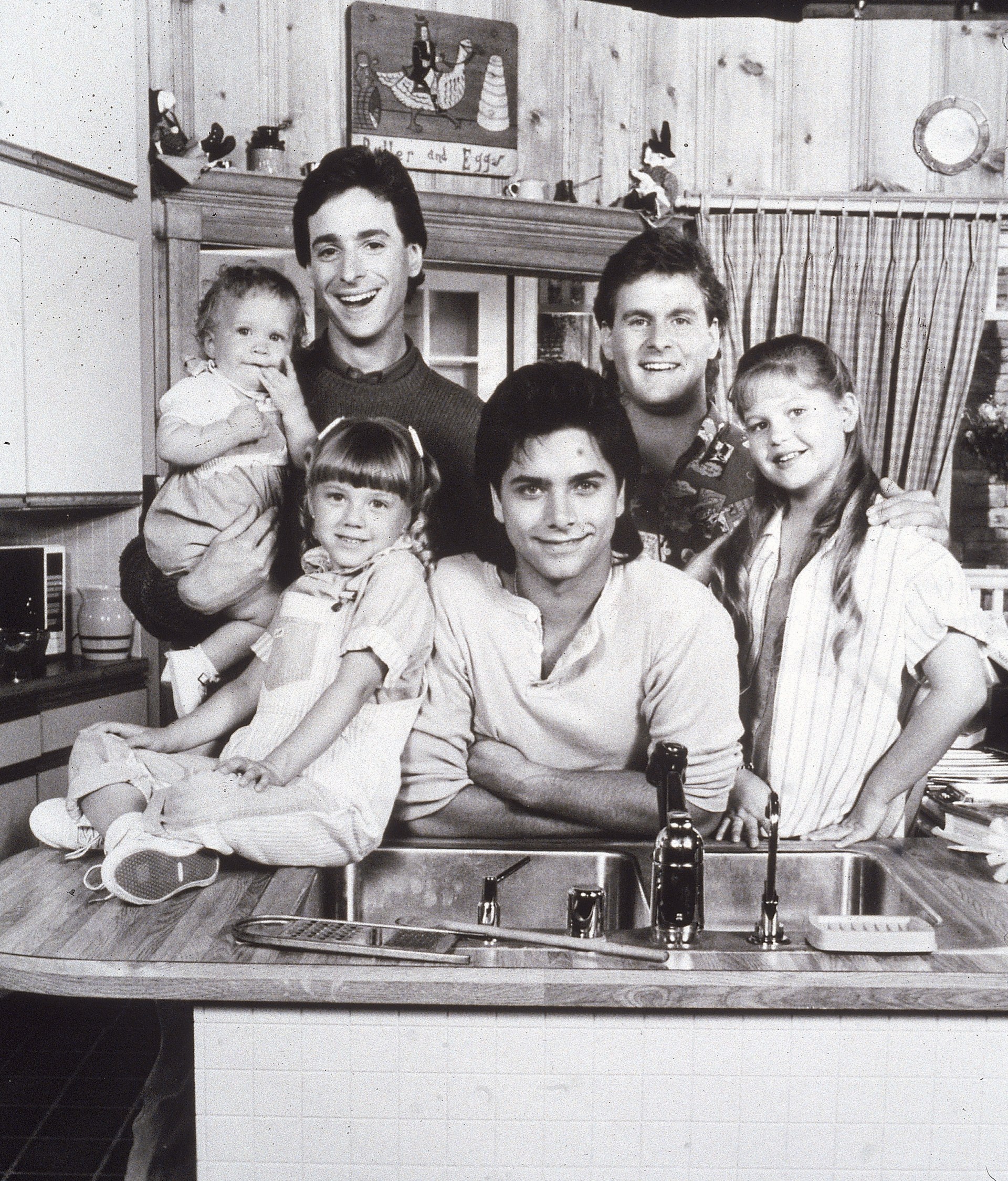 Black-and-white promo photo of the Full House cast posing around a kitchen sink, including two adult men, one young man, two girls, and a baby, circa 1987.