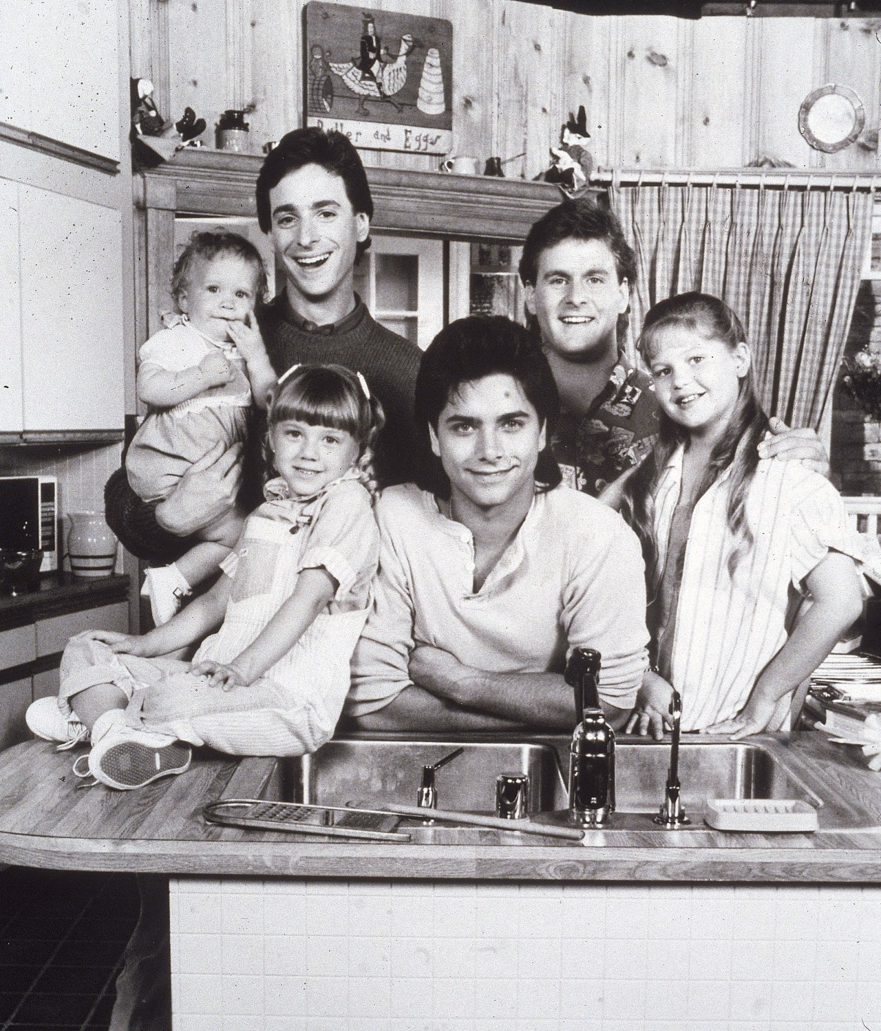Black-and-white promo photo of the Full House cast posing around a kitchen counter, including two adult men, a young man, two girls, and a toddler.