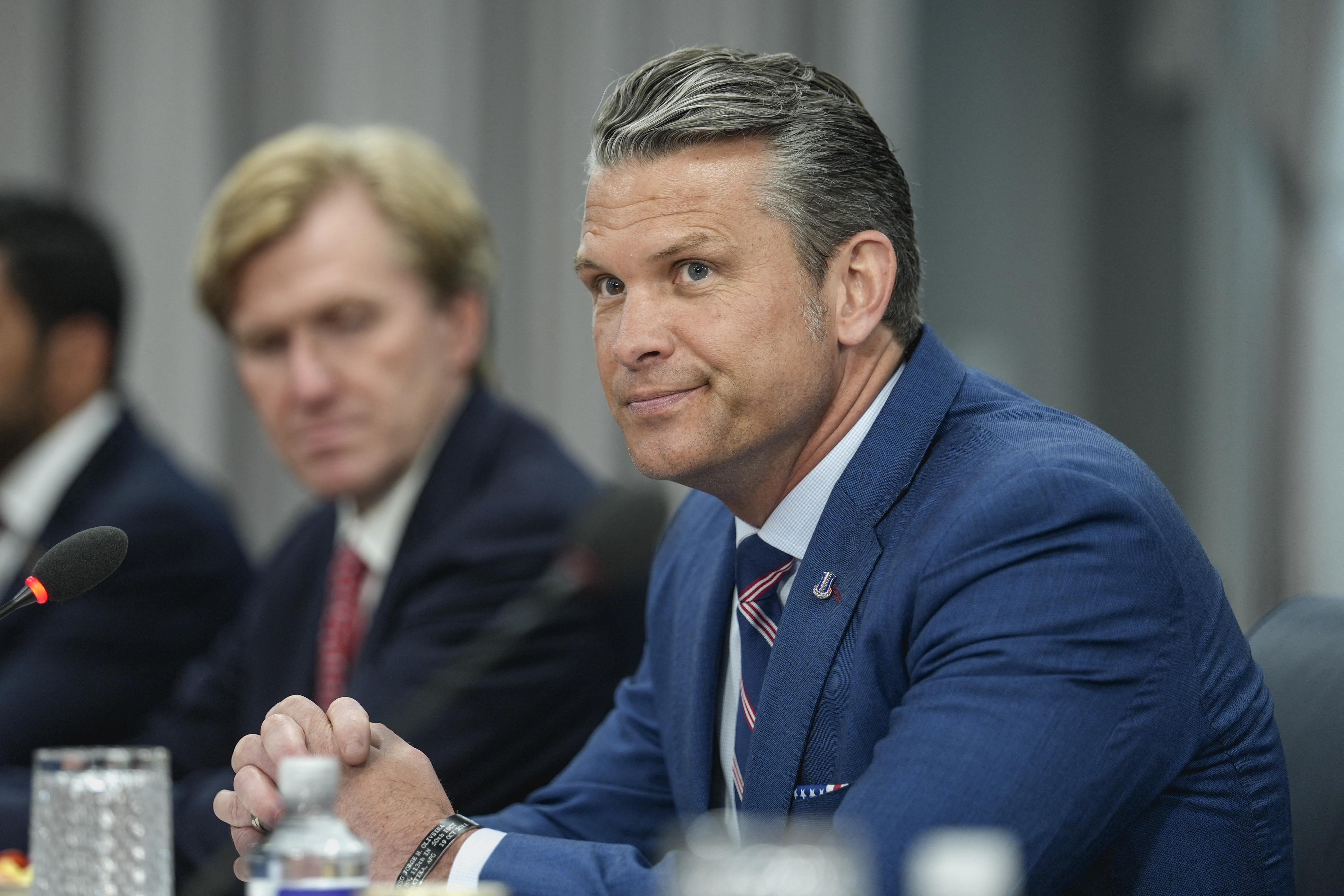 Pete Hegseth in a blue suit and patriotic tie sits at a conference table with hands clasped, a microphone nearby and another official blurred in the background.