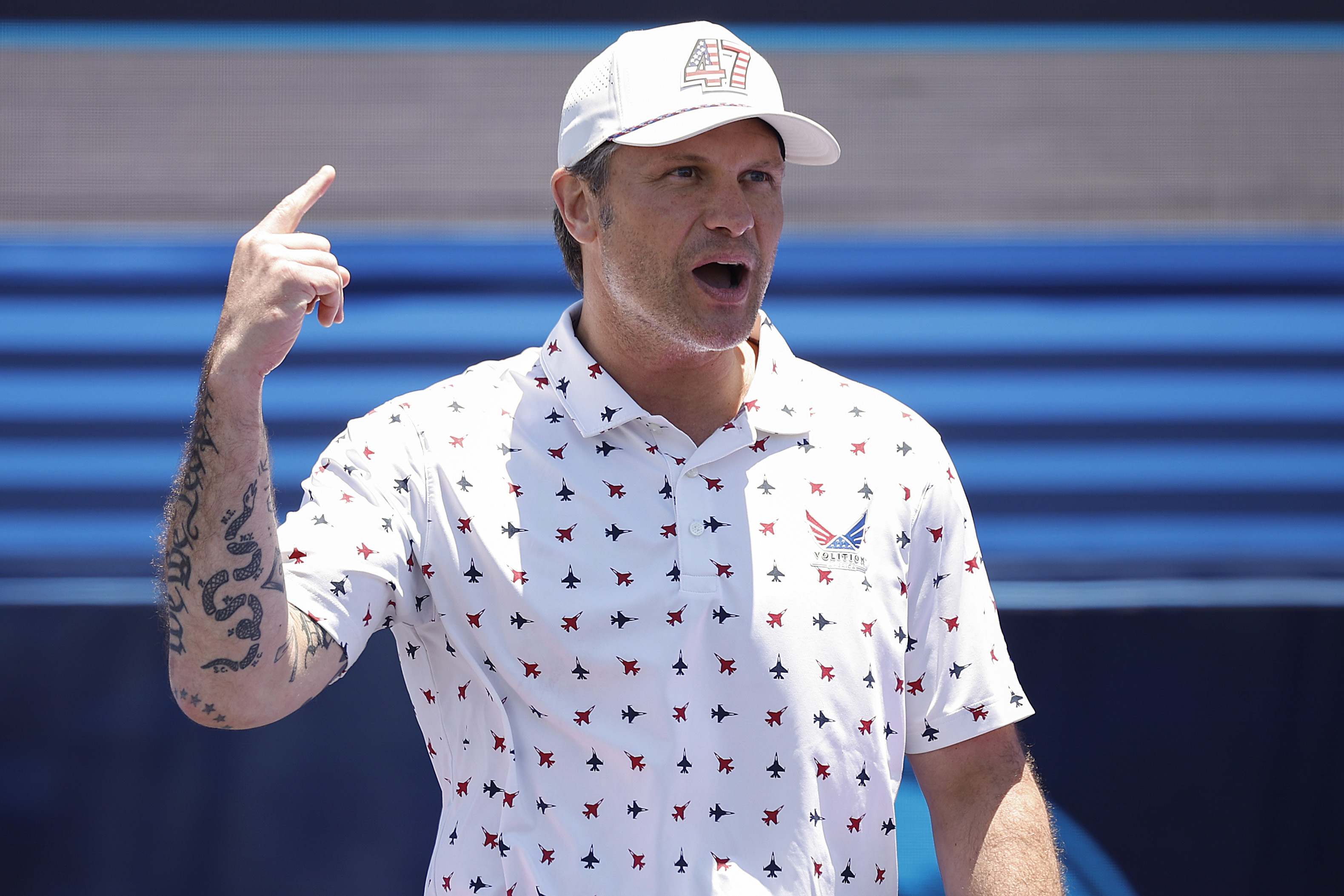 Pete Hegseth in a white jet-print polo and '47 cap points one finger upward while speaking at a NASCAR pre-race ceremony.