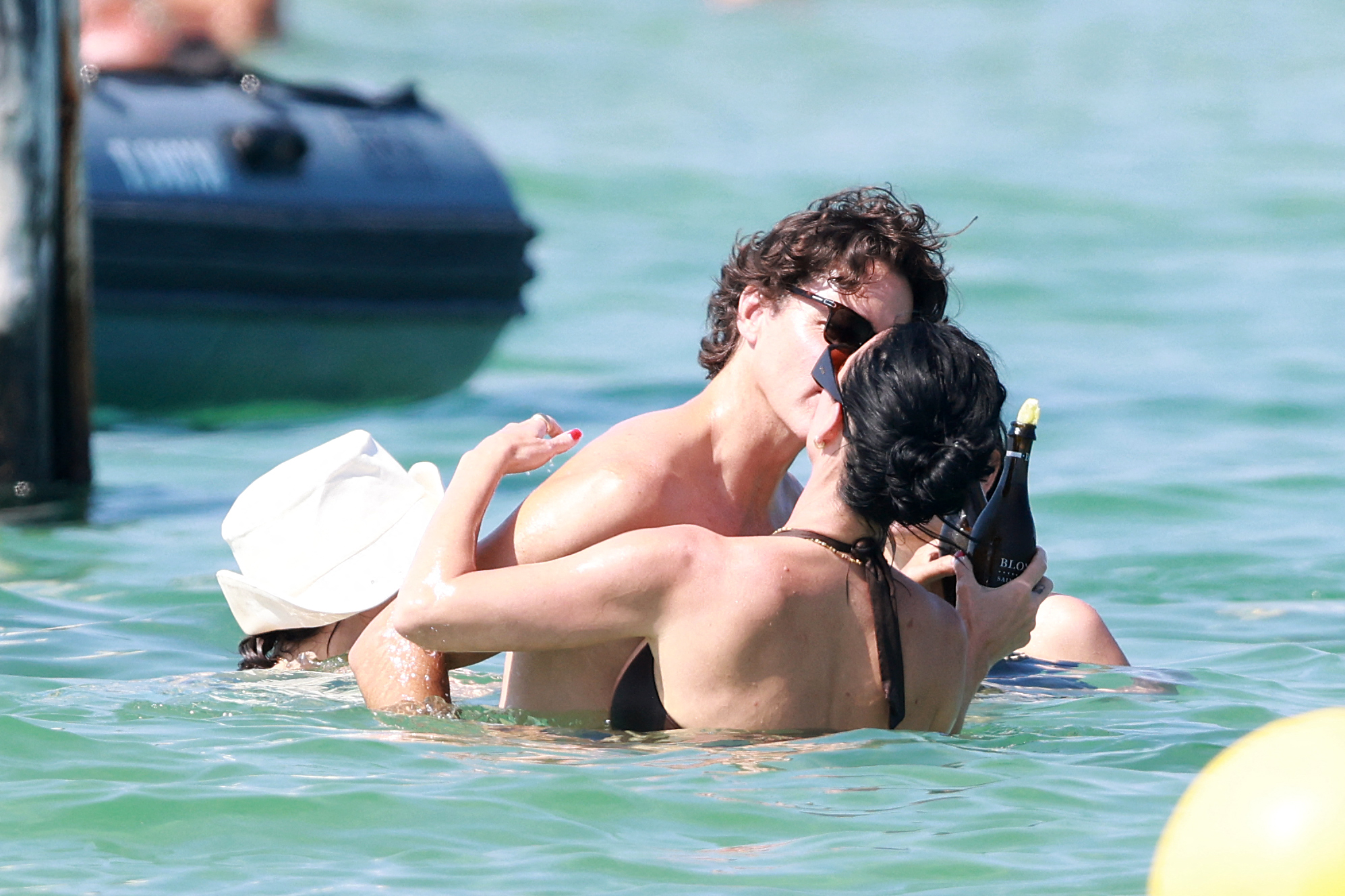 A shirtless man and a woman in a brown bikini kiss while standing in the sea, the woman holding a dark bottle of sparkling wine.