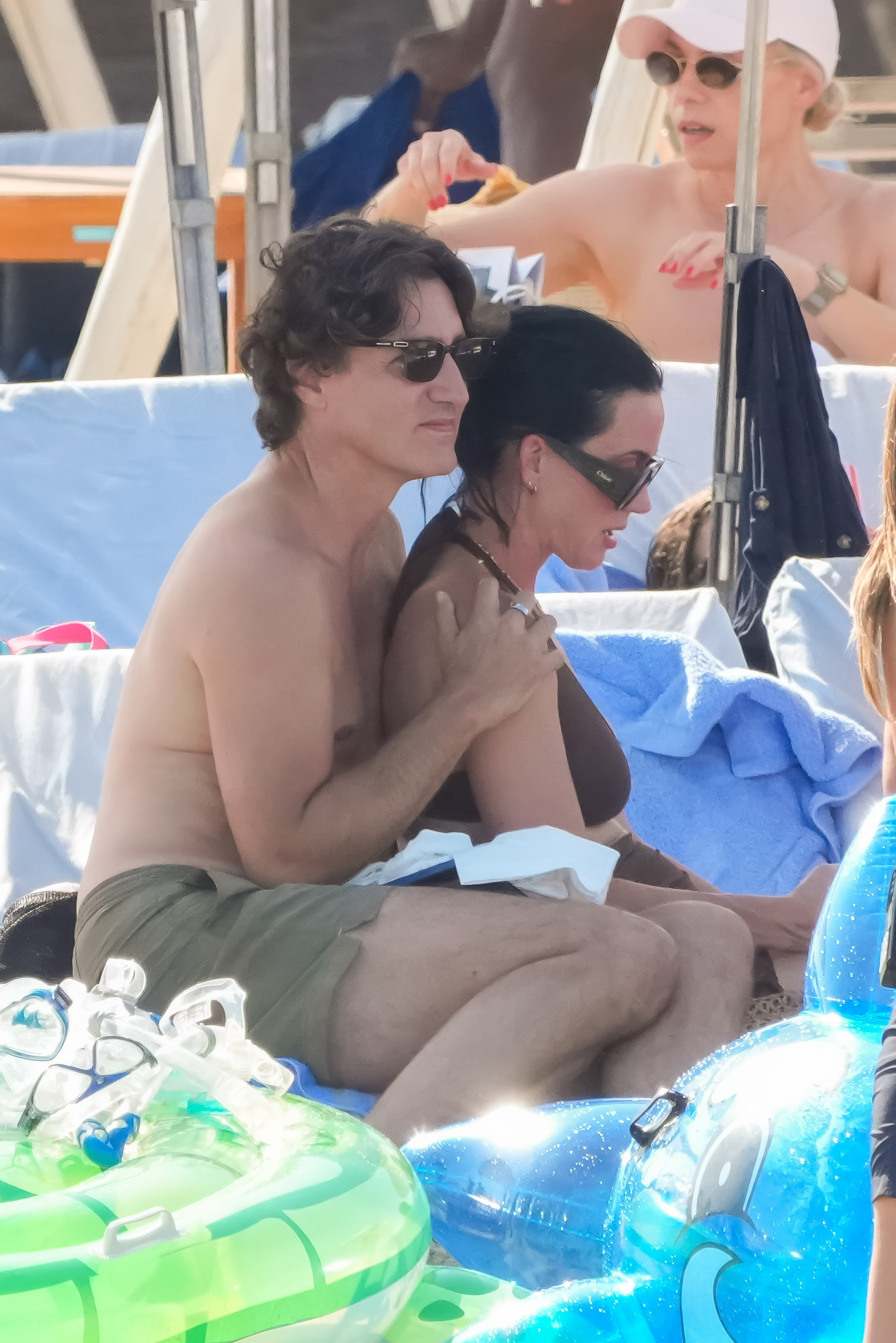 A shirtless man with curly hair and sunglasses sits closely behind a dark-haired woman in a brown bikini top on a beach lounger, surrounded by pool floats and snorkeling gear.