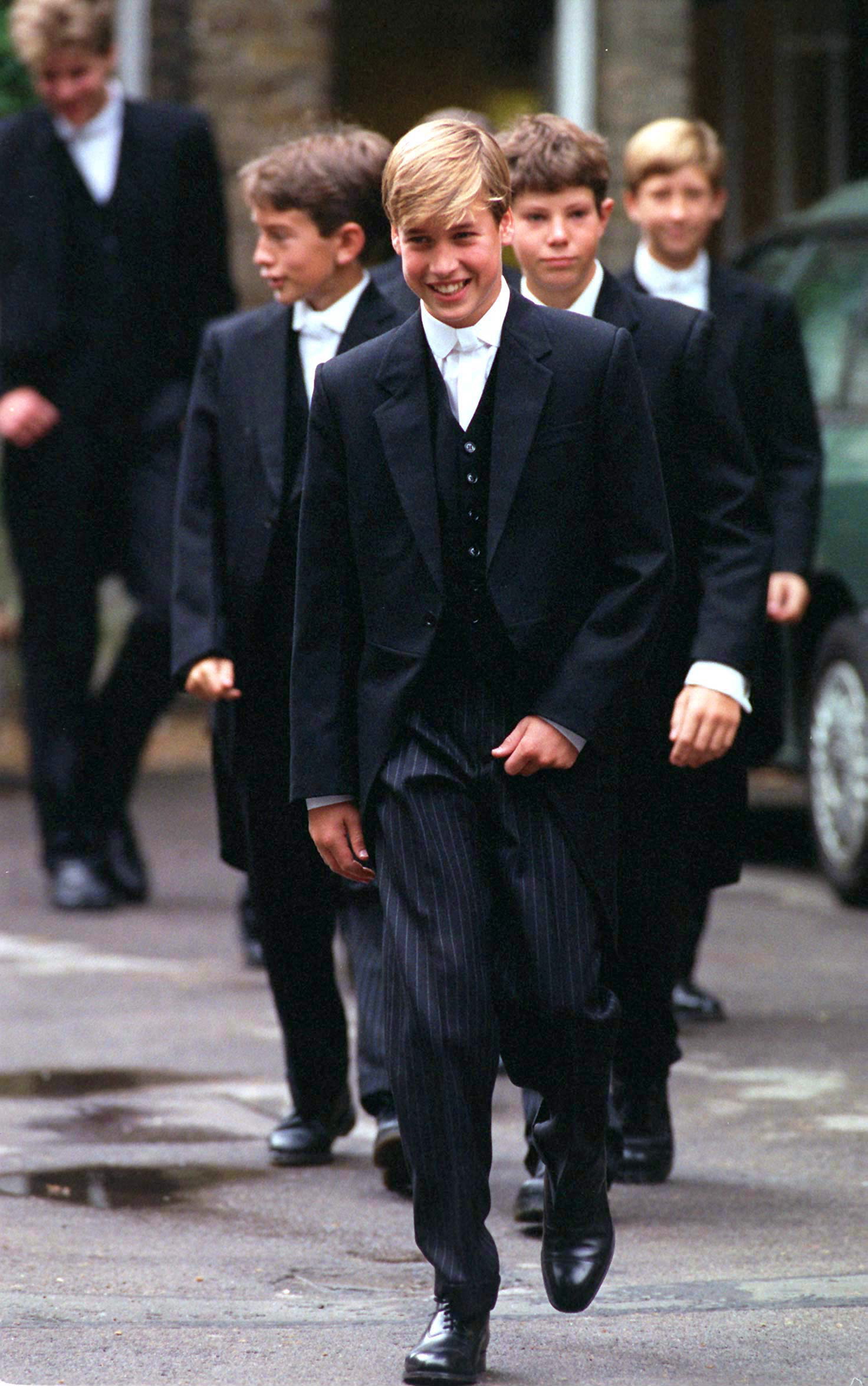 Smiling blonde teenage boy in Eton morning suit walking ahead of a group of boys in matching formal uniforms on a wet stone path.