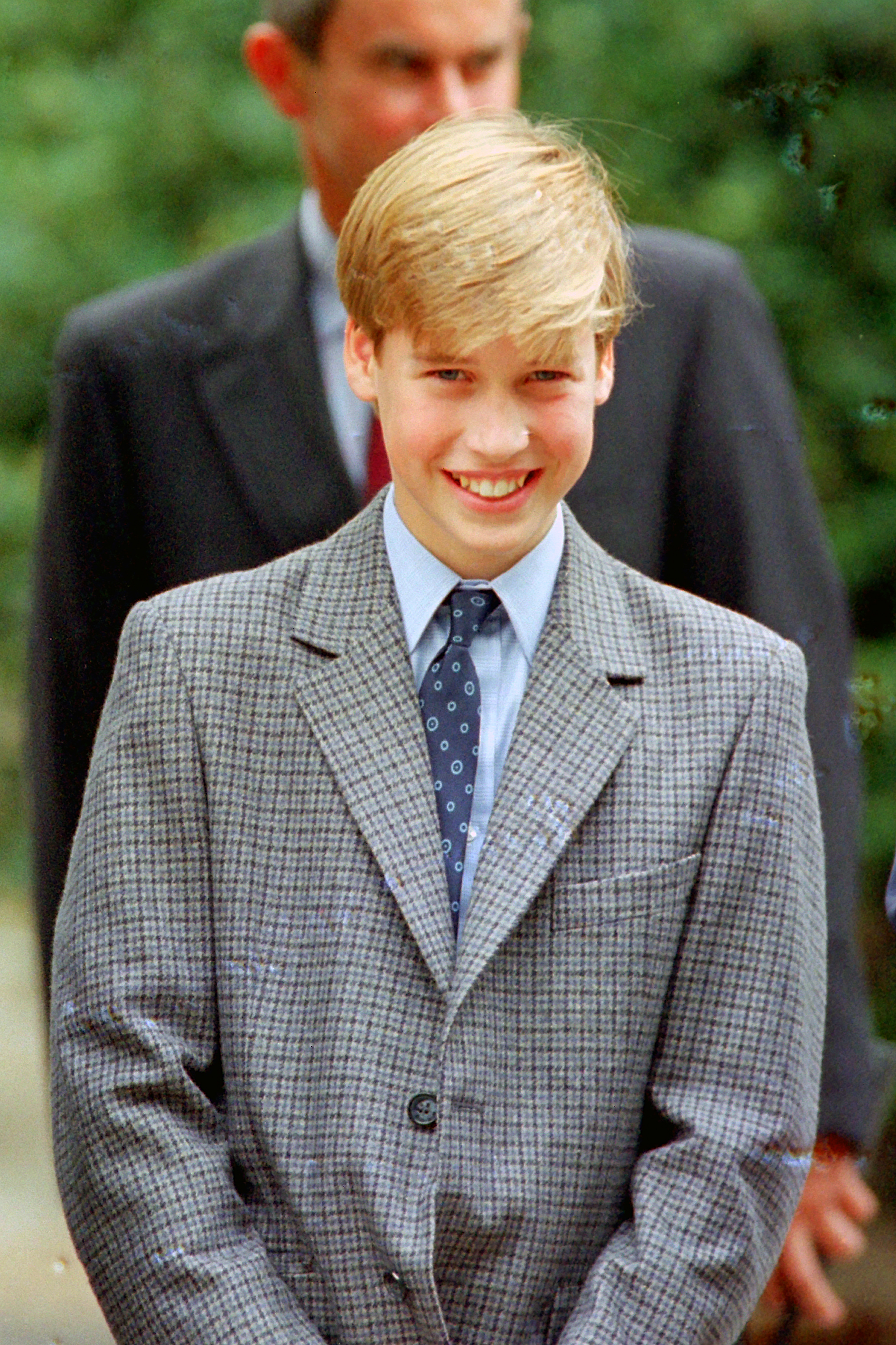 Young teenage boy with blonde hair in a grey checked blazer and navy tie, smiling outdoors with a man in a dark suit blurred behind him.