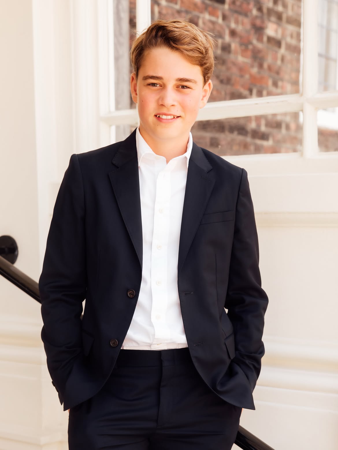 Teenage boy in a navy blazer and white shirt, hands in pockets, smiling in front of a brick wall and white-framed window.