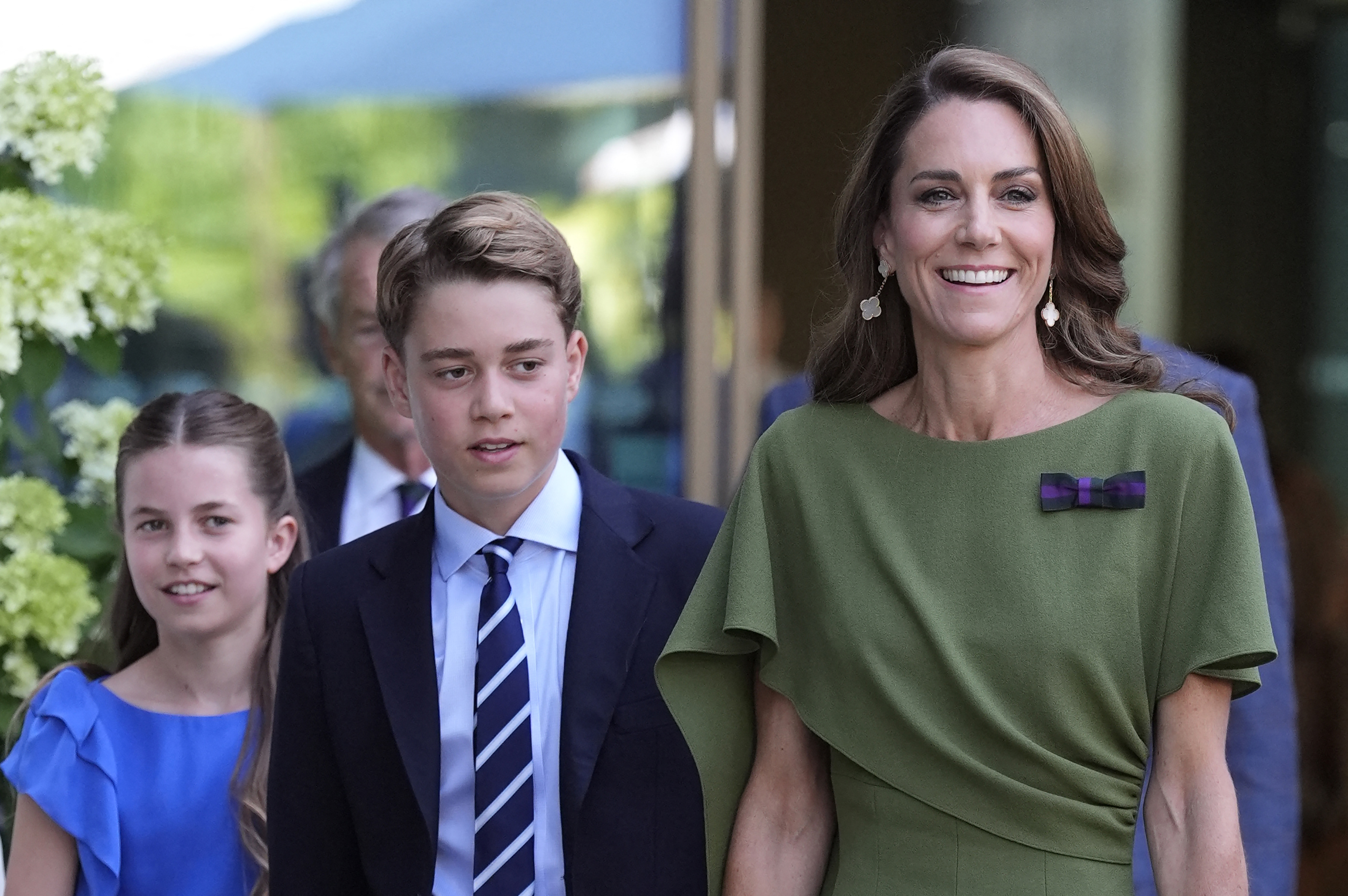 Woman in an olive green dress walks alongside a teenage boy in a navy suit and tie and a girl in a blue dress at Wimbledon.