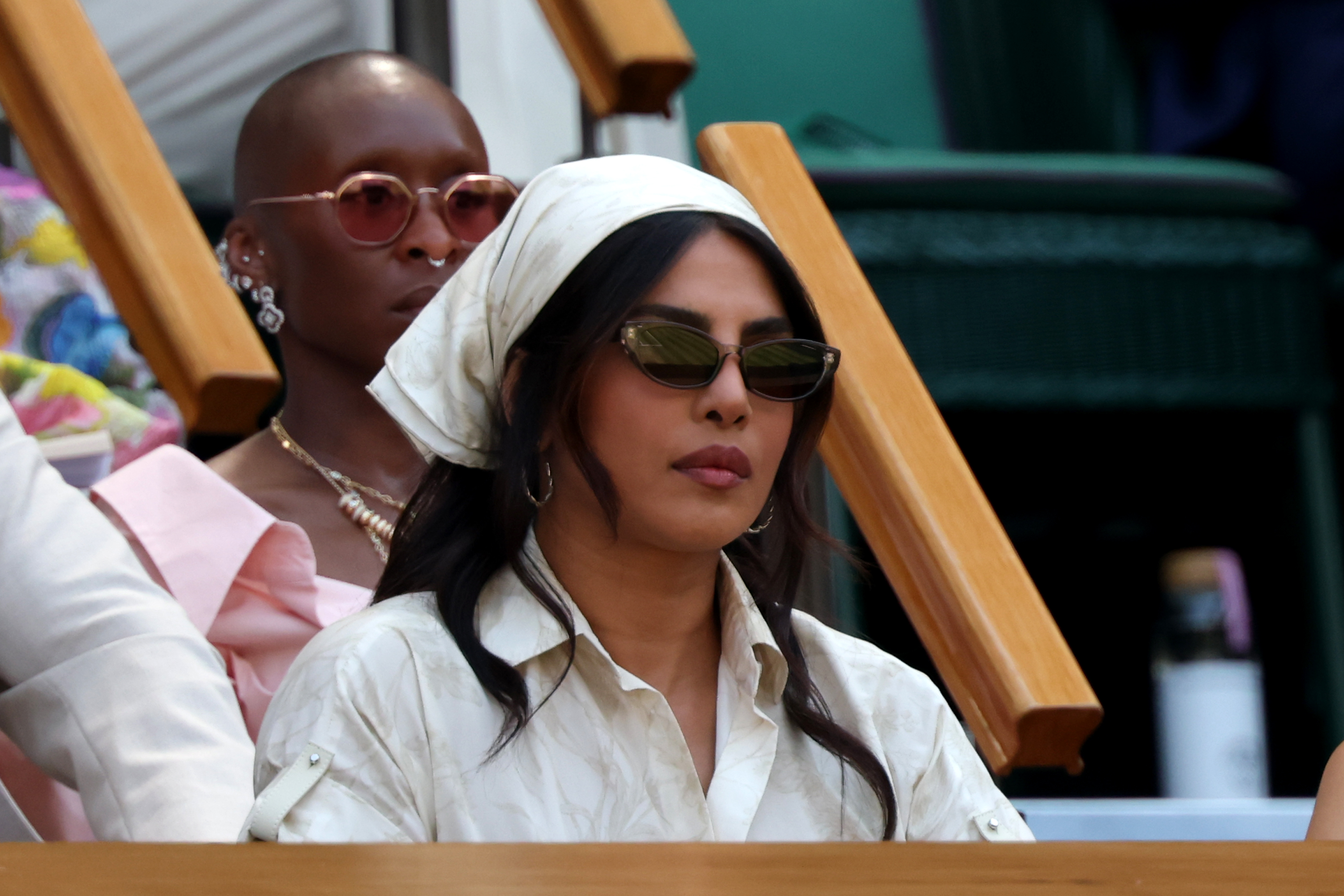 Priyanka Chopra Jonas seated in Wimbledon's Royal Box wearing a cream headscarf and small dark sunglasses, with another woman in pink behind her.