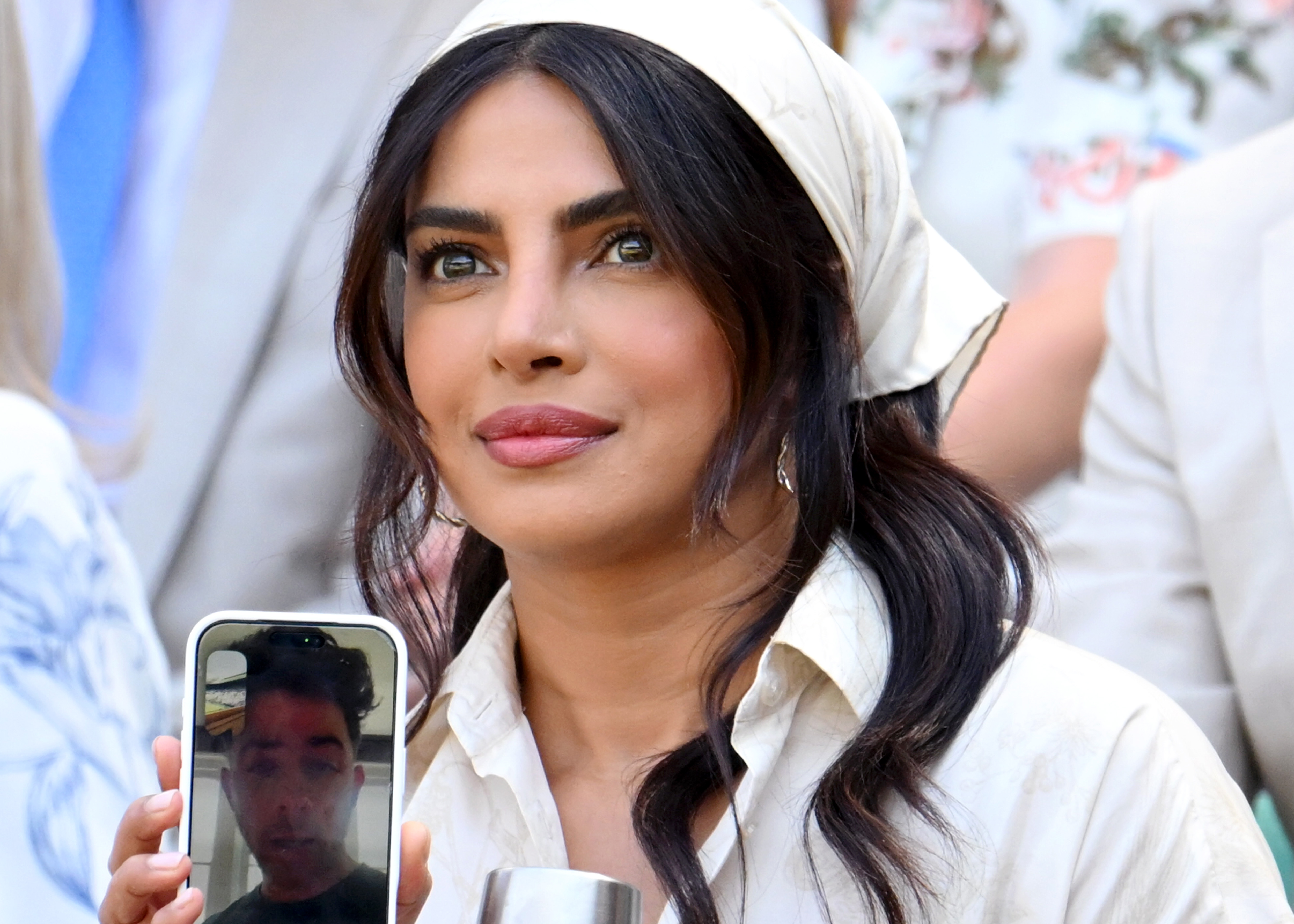 Woman with dark hair and a cream headscarf holds up a phone showing a man on FaceTime while holding a silver cup, at Wimbledon.
