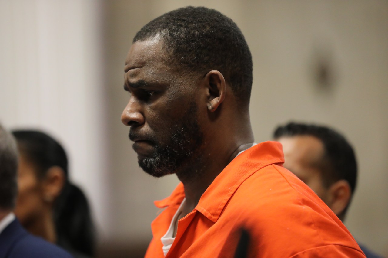 Man in orange jail jumpsuit standing in profile at a court hearing, with other individuals blurred in the background.