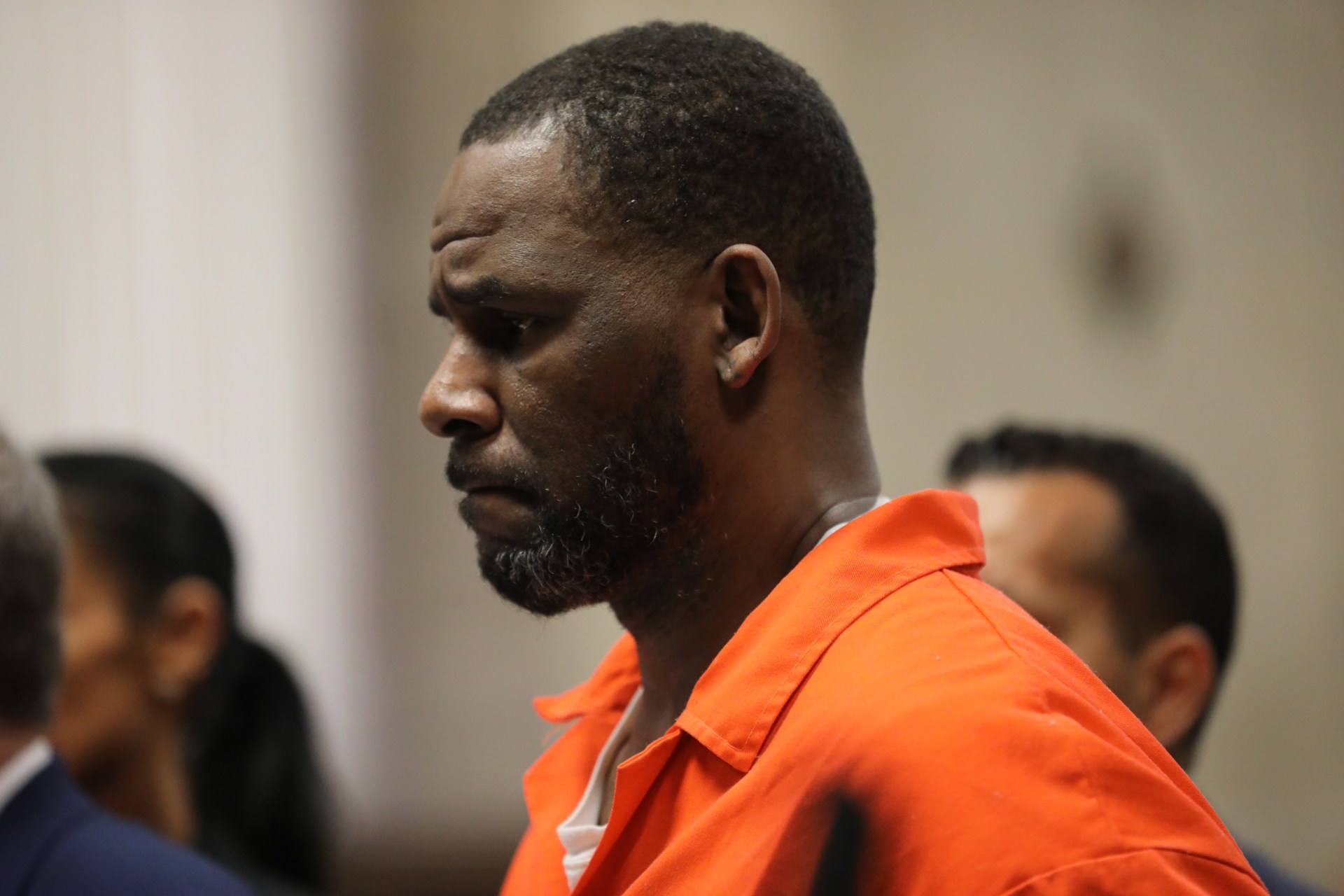 Man in orange jail jumpsuit standing in profile at a court hearing, with other individuals blurred in the background.