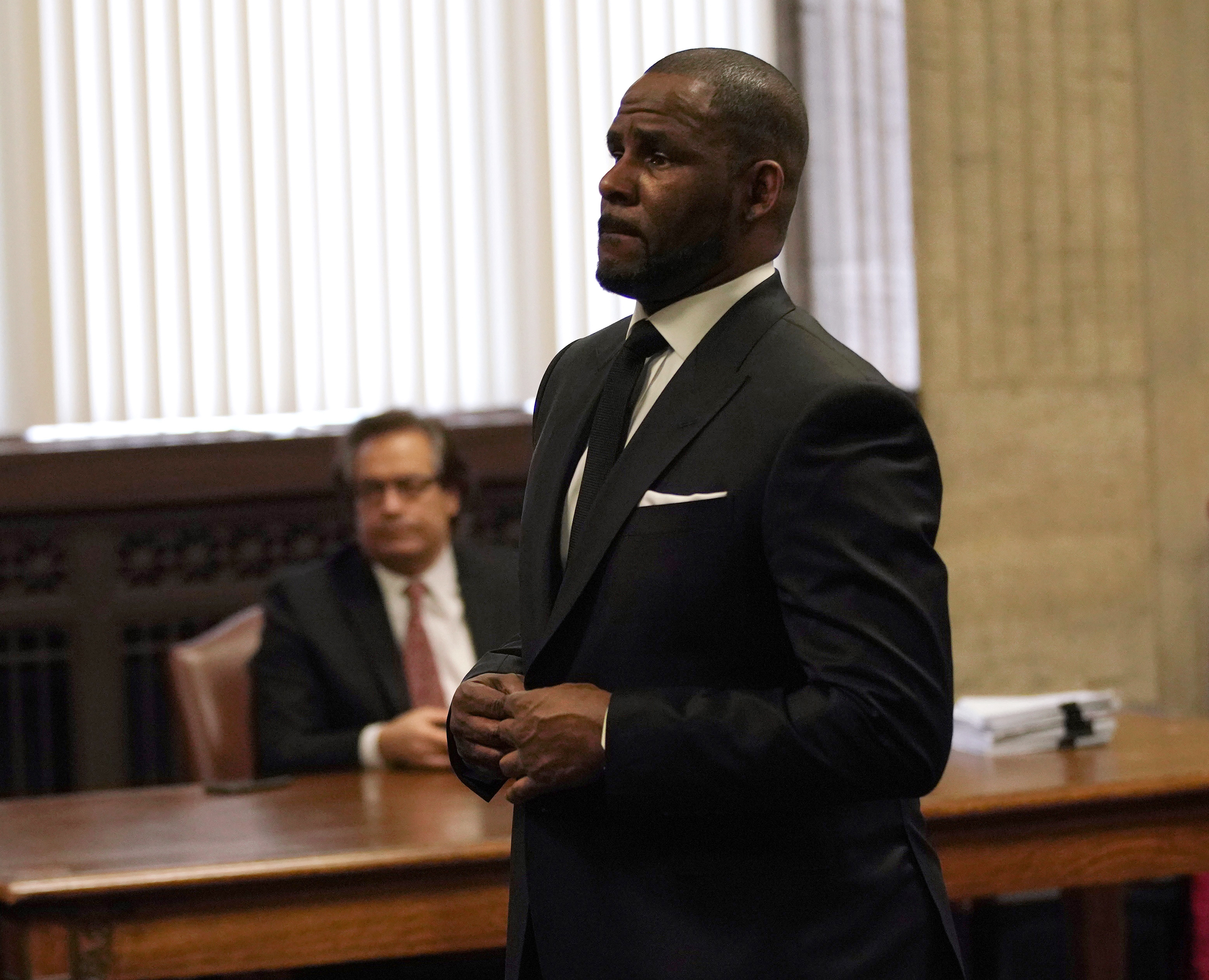 Man in a black suit with hands clasped standing in a wood-paneled courtroom, with a seated man in a suit visible behind him.