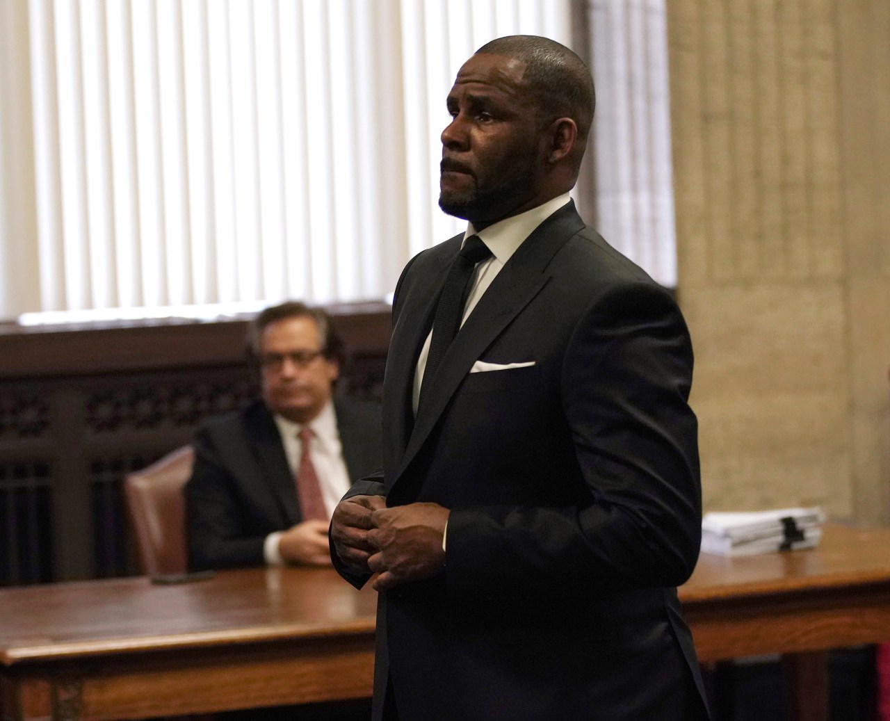 Man in a black suit with hands clasped standing in a wood-paneled courtroom, with a seated man in a suit visible behind him.