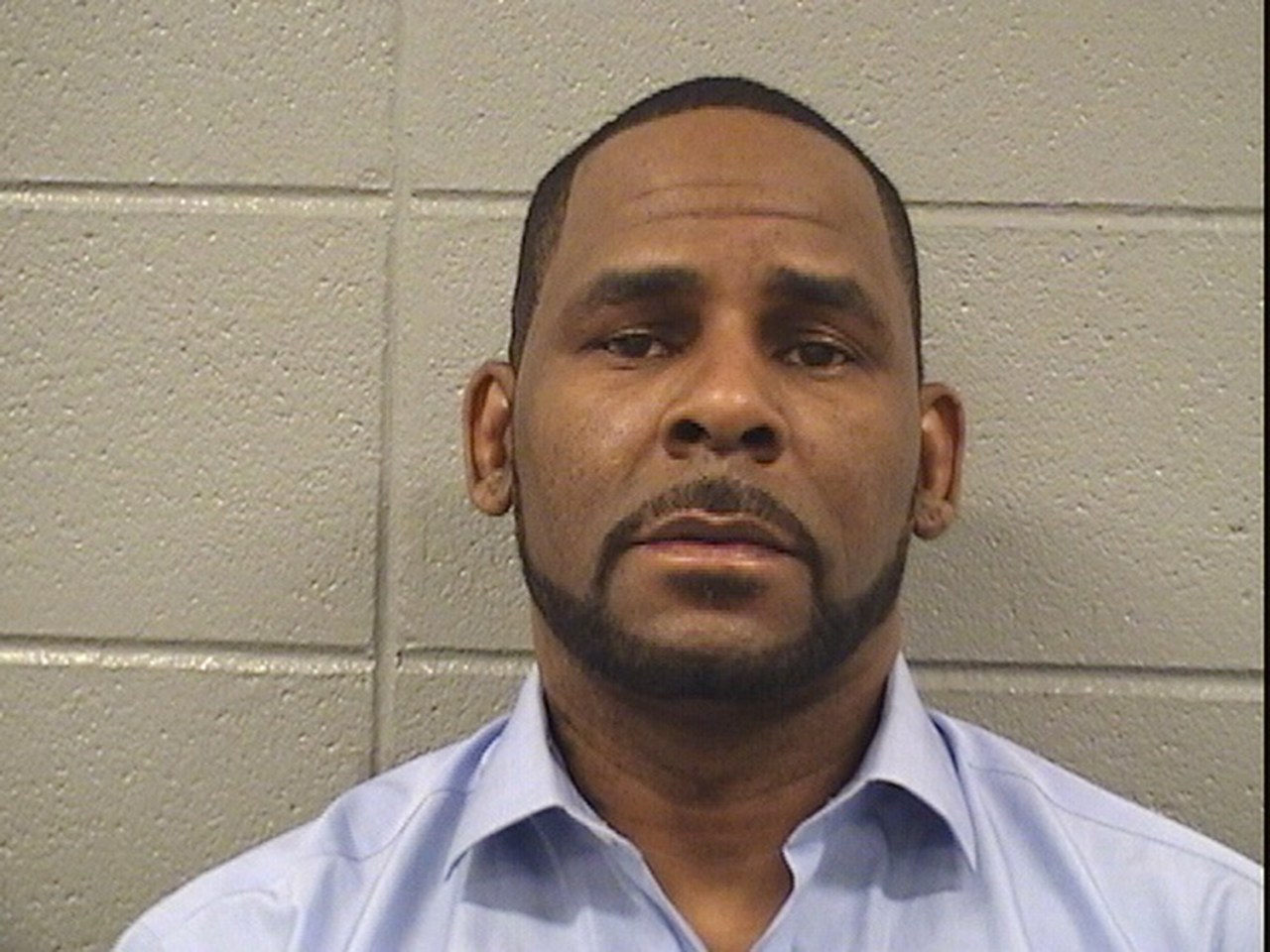 Close-up booking mugshot of a man in a light blue shirt against a concrete block wall.