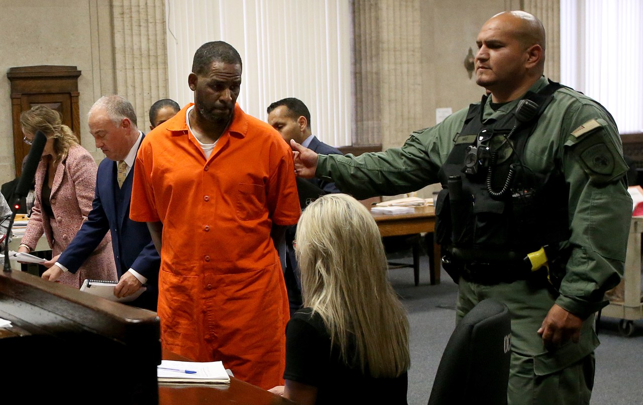 Man in orange prison jumpsuit escorted by a uniformed officer inside a courtroom, with attorneys visible in the background.