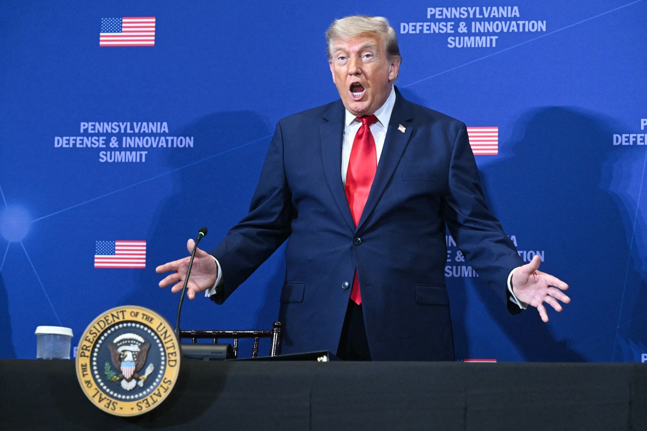 Donald Trump standing with arms outstretched and mouth open in front of a blue Pennsylvania Defense and Innovation Summit backdrop, with the presidential seal on the table in front of him.