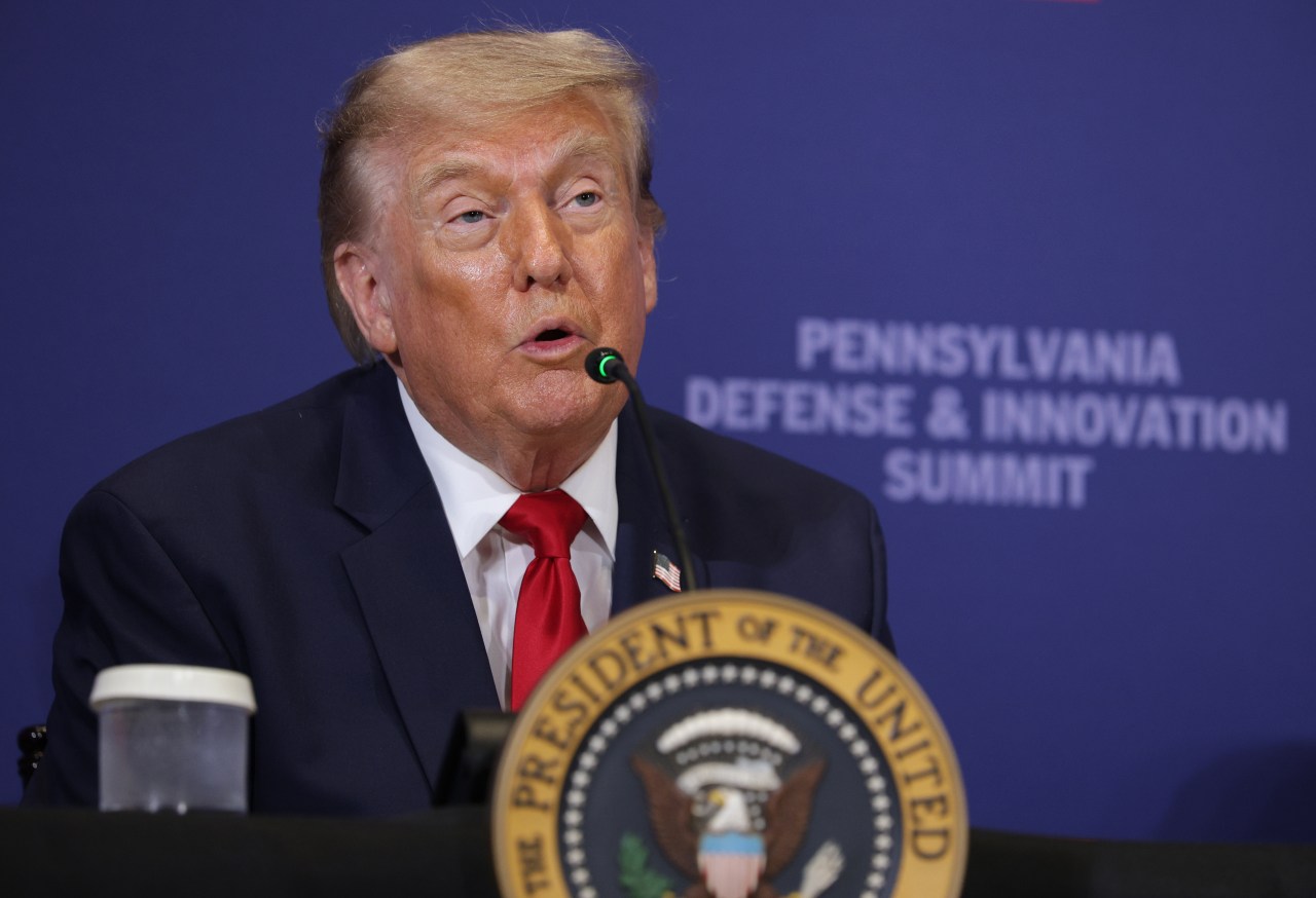 Donald Trump speaking at a microphone at a podium bearing the presidential seal, with a blue Pennsylvania Defense and Innovation Summit backdrop behind him.