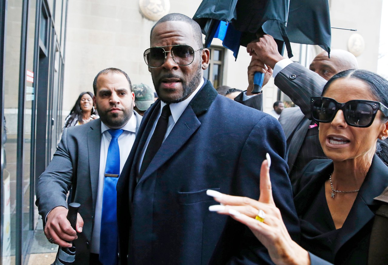Man in a navy overcoat and aviator sunglasses exiting a courthouse amid a crowd of people.