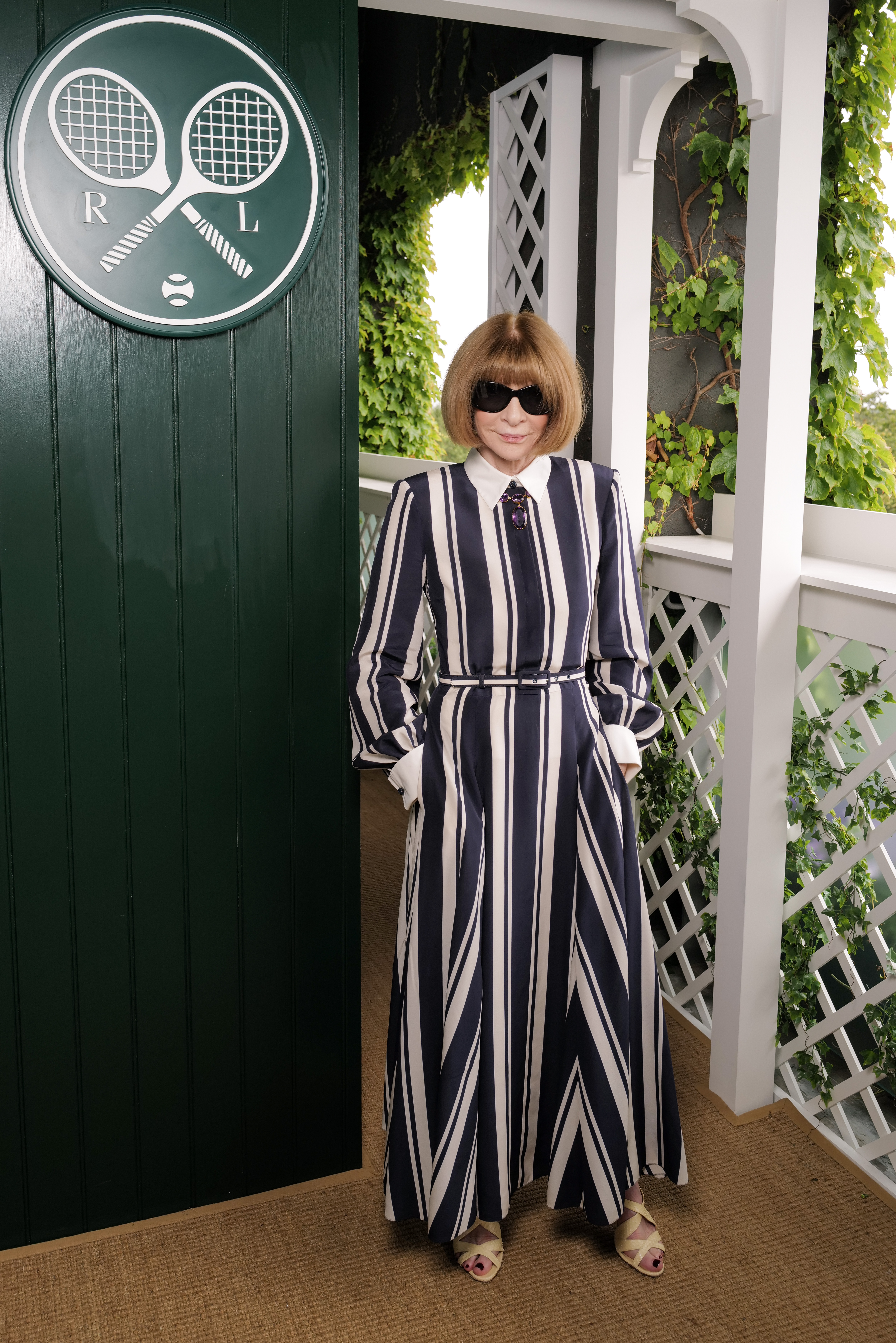 Woman with a blonde bob and black sunglasses wearing a navy and white striped floor-length dress, standing at the Ralph Lauren Wimbledon suite.