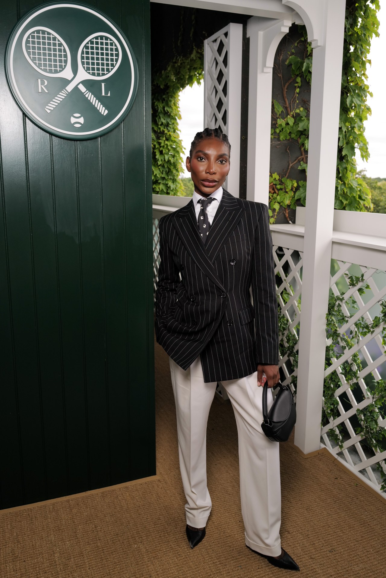 Michaela Coel in a black pinstripe blazer, white trousers, and black heels, posing beside a green Ralph Lauren Wimbledon door.