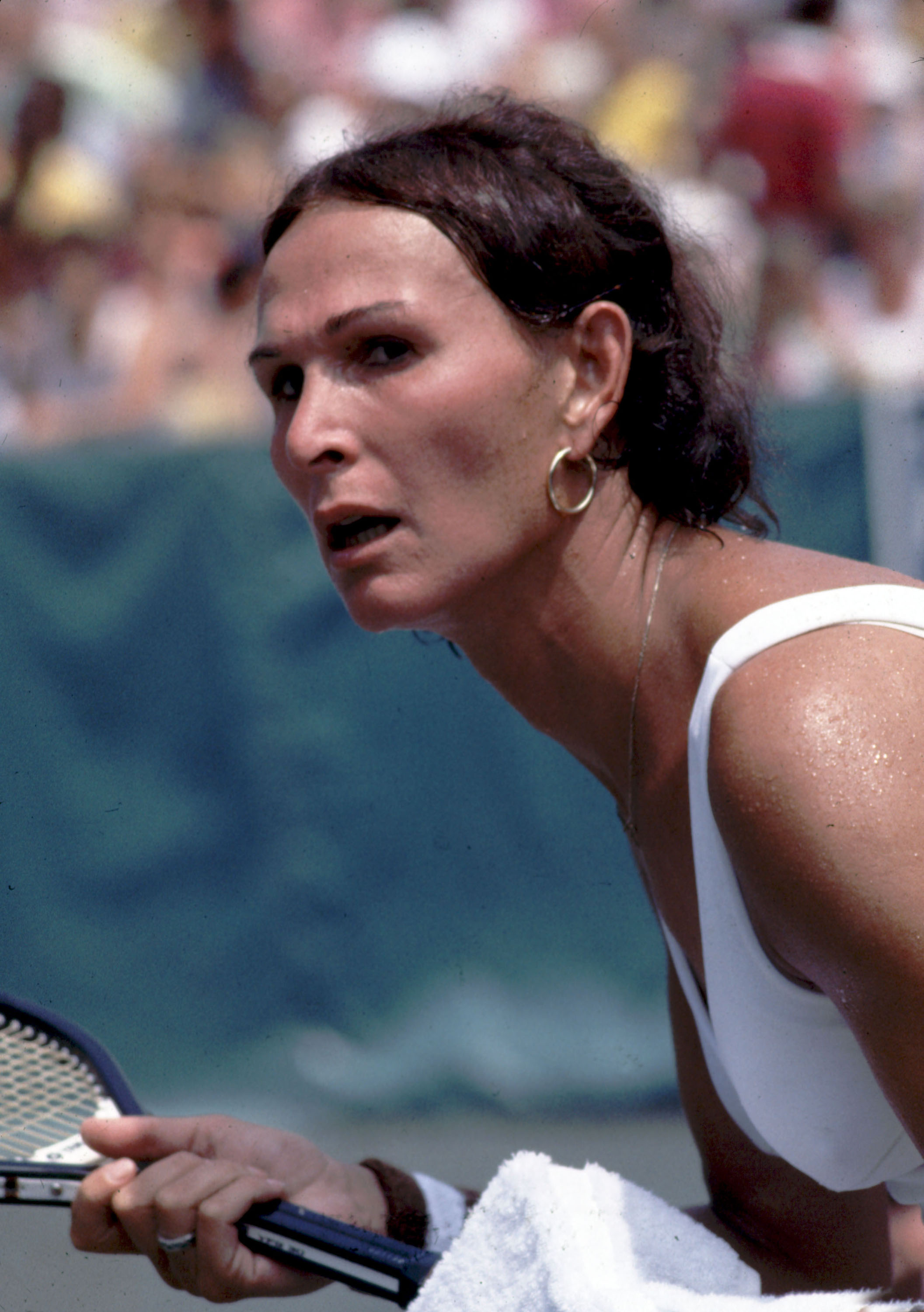 Close-up of a female tennis player in a white sleeveless top, leaning forward with mouth open and gripping a racket, crowd blurred in background.