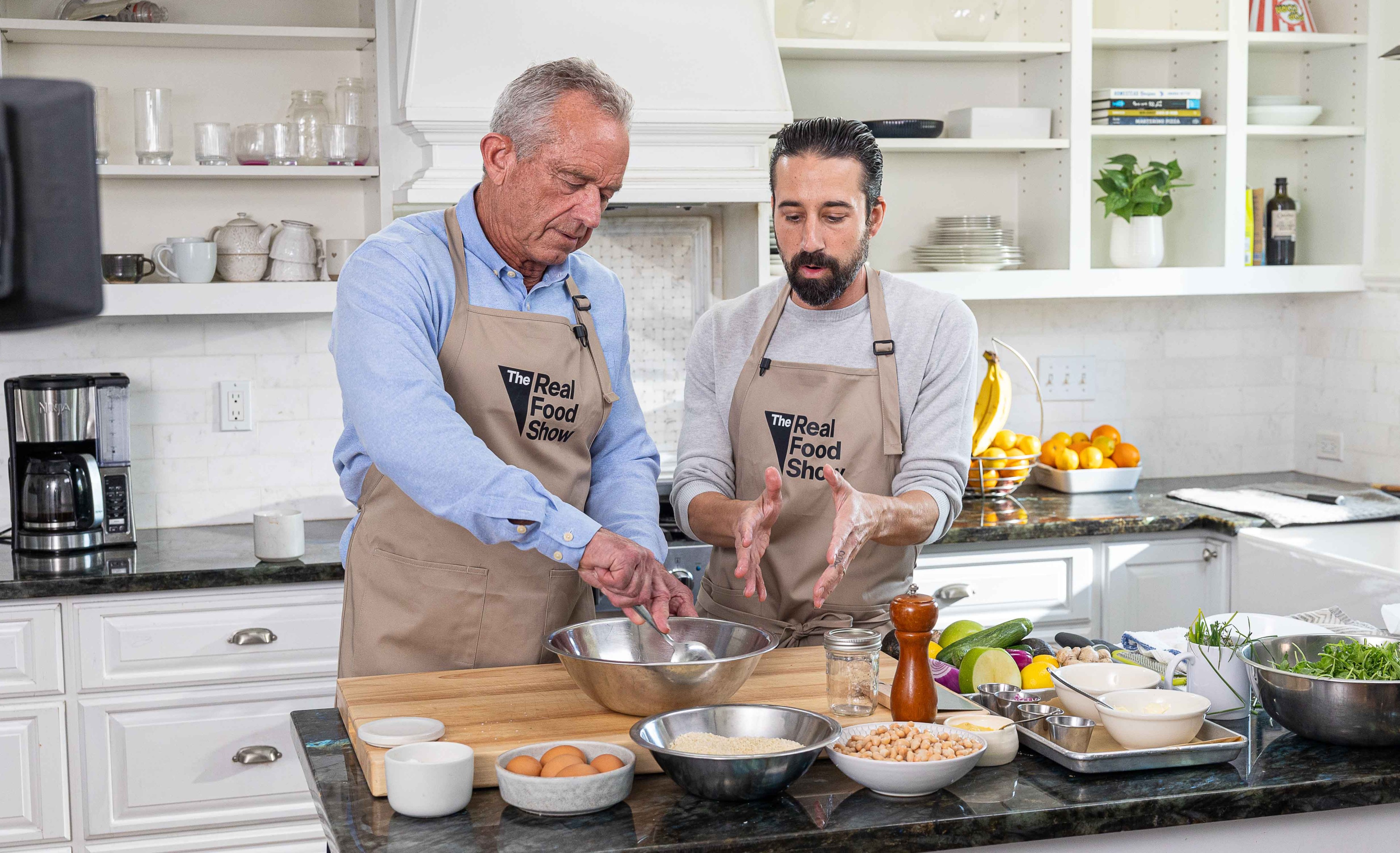 Two men in 'The Real Food Show' aprons work together at a kitchen island, one stirring a metal mixing bowl while the other gestures with his hands.