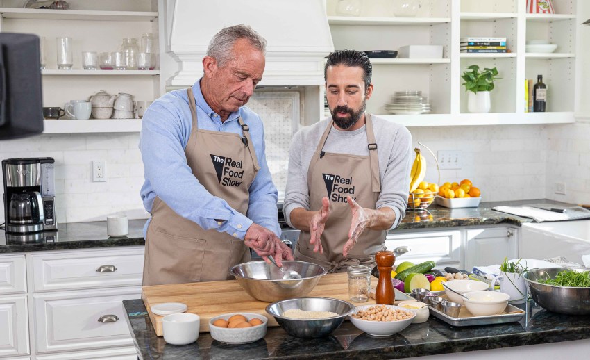 Two men in 'The Real Food Show' aprons work together at a kitchen island, one stirring a metal mixing bowl while the other gestures with his hands.