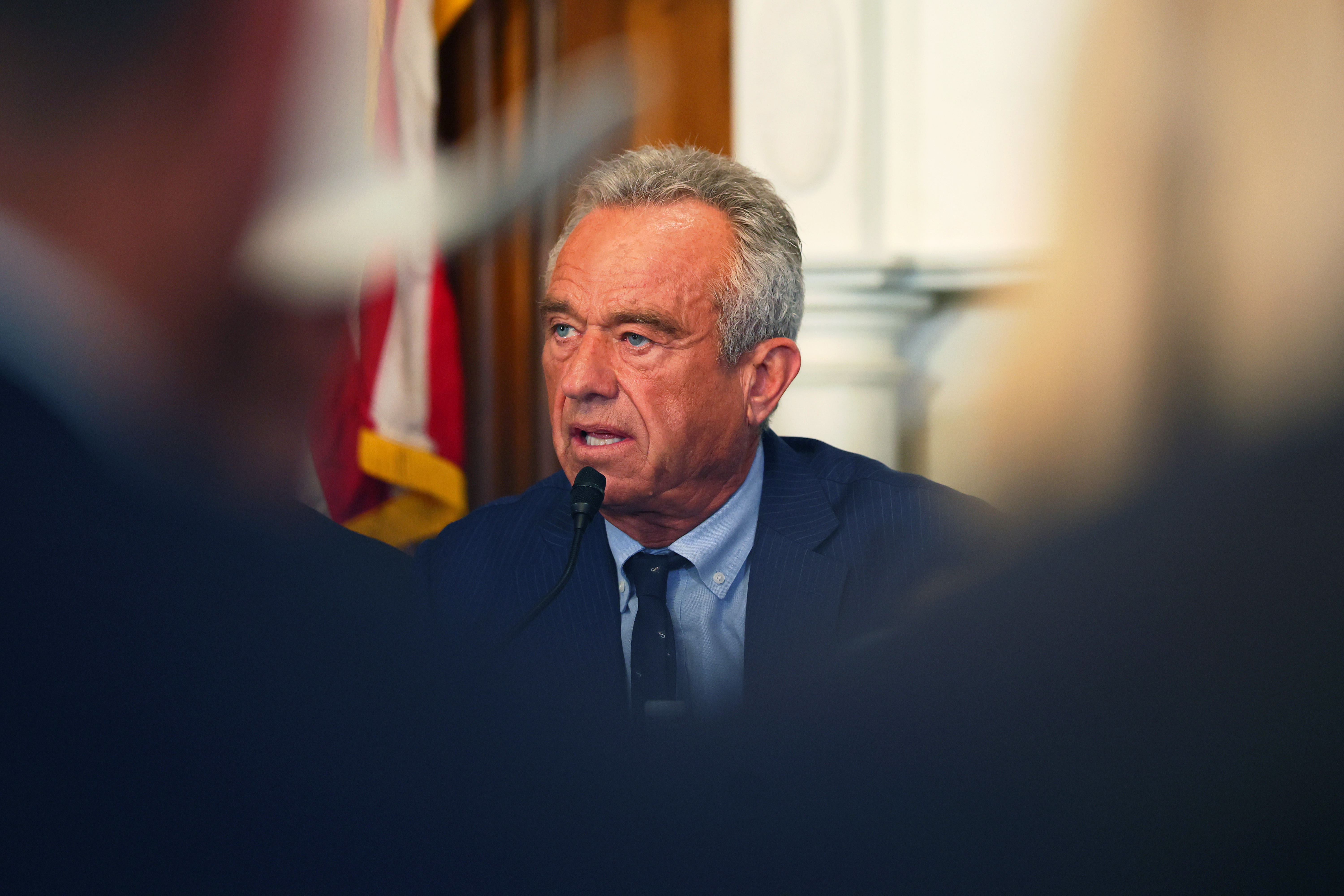 Man in navy suit speaking into a microphone at a roundtable, with a U.S. flag visible in the background.