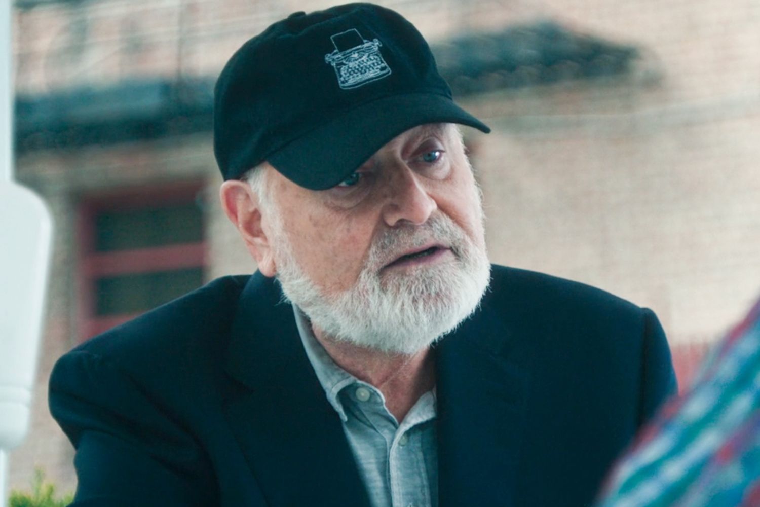 Older man with white beard wearing a black cap with a typewriter logo and dark blazer, looking off to the side in a scene from The Bear.