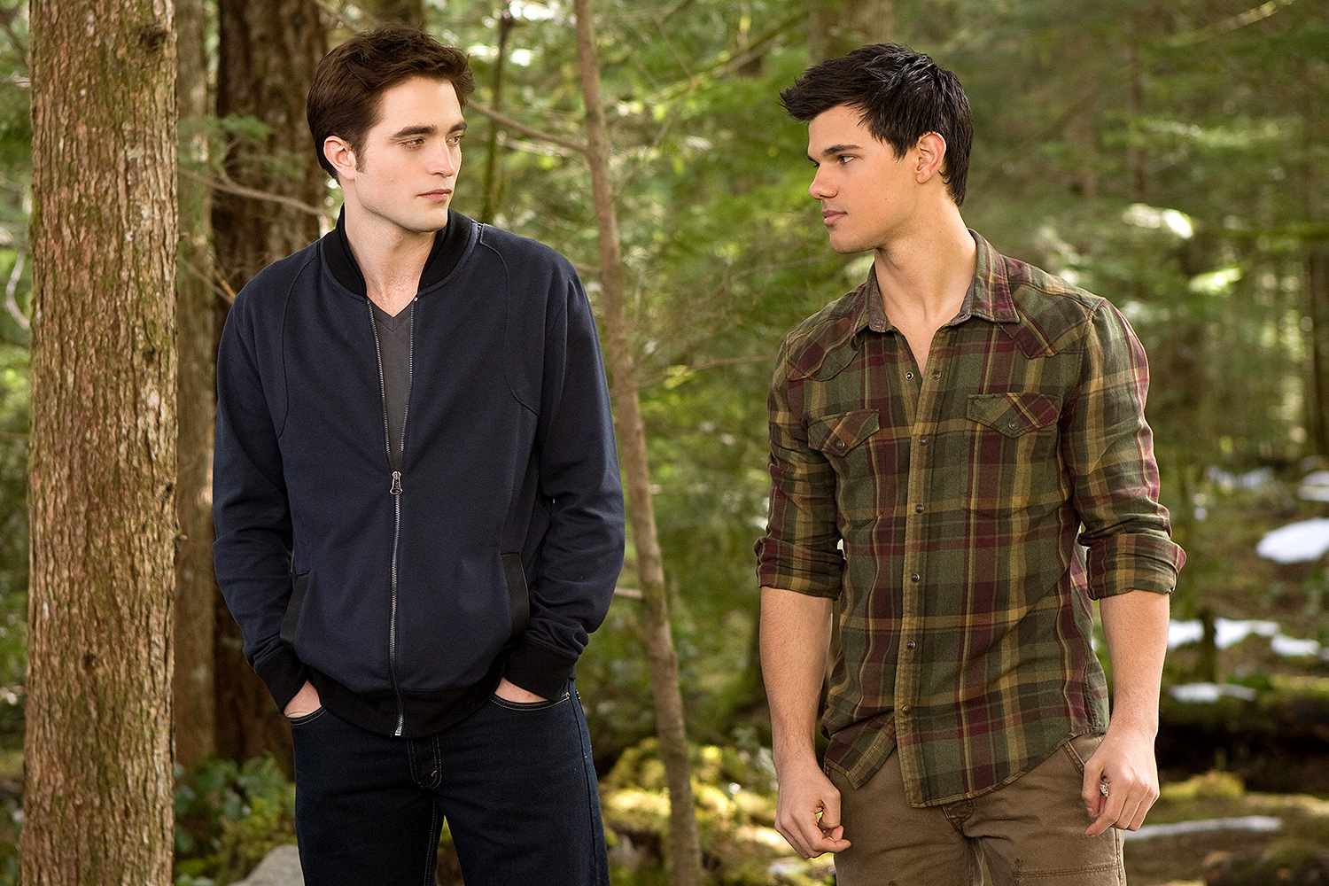 Two young men — one in a navy zip-up jacket, one in a green plaid shirt — stand face to face in a forested outdoor setting.