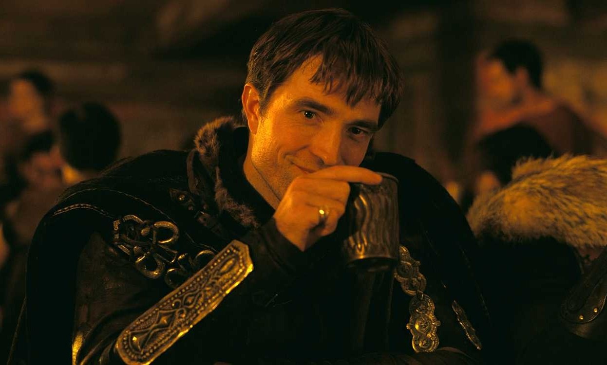 A man in dark medieval armor and fur-trimmed robes smirks while raising an ornate metal cup at a dimly lit banquet hall.