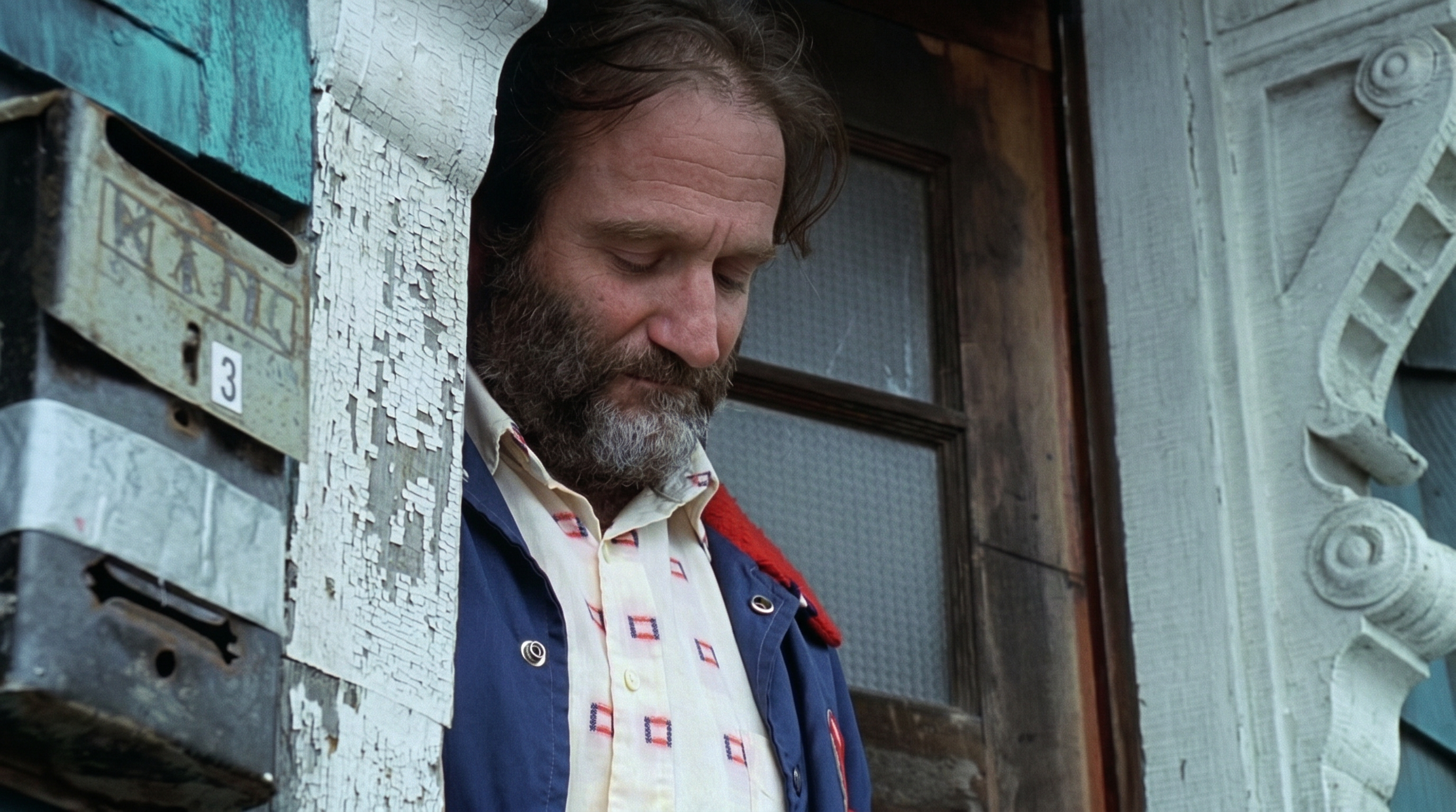 A bearded man in a blue jacket leans against a weathered doorframe with peeling paint and old mailboxes, looking downward.