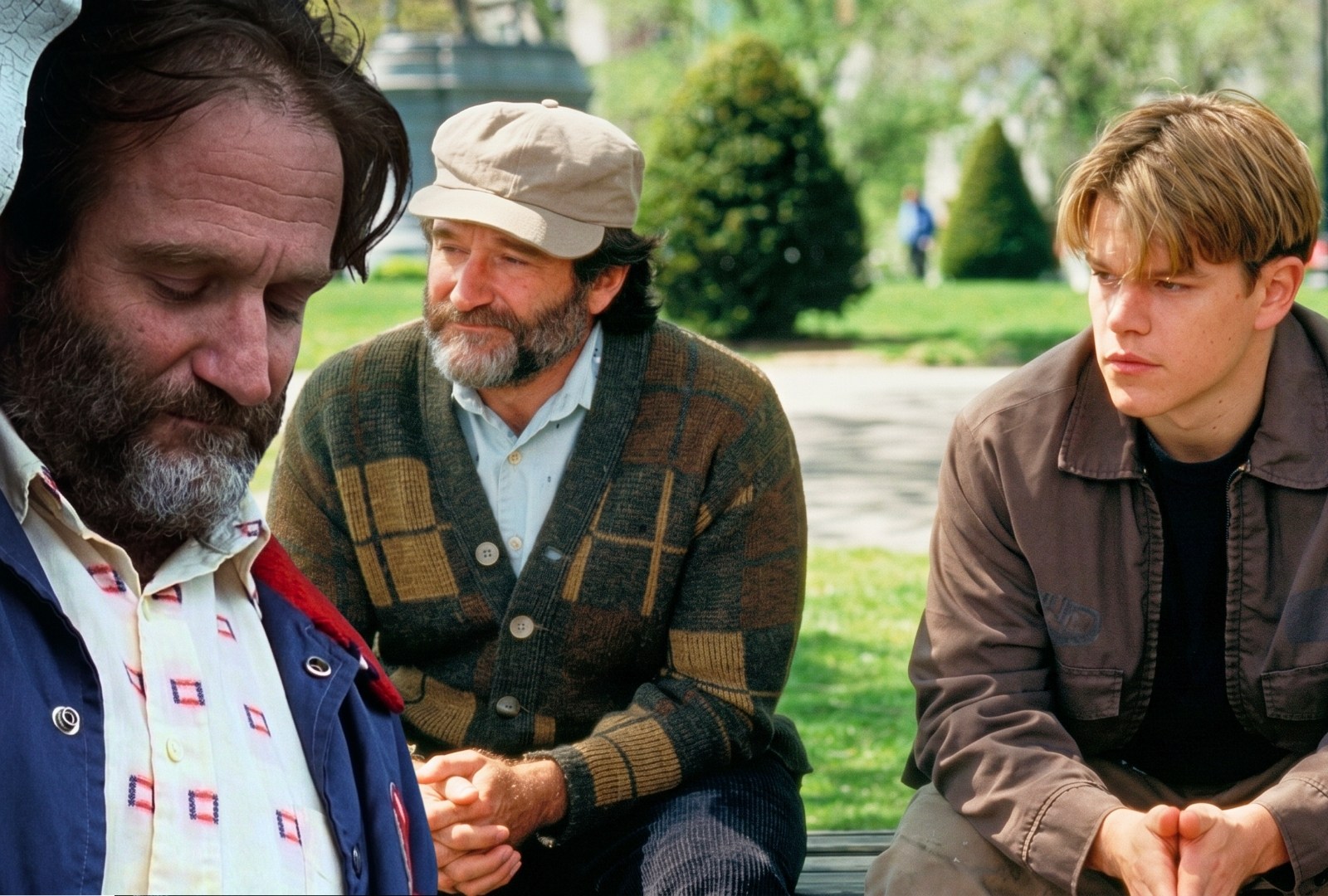 Two men sitting together on a park bench in a green park, one older wearing a newsboy cap and patterned cardigan, one younger in a brown jacket.