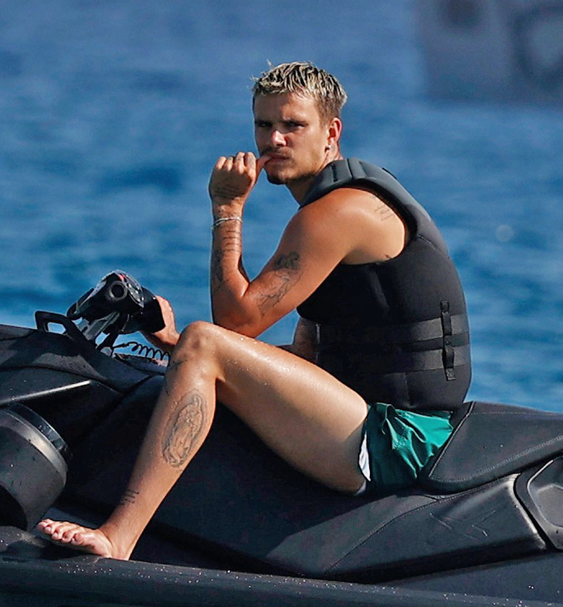 Young blond man in a black life vest and teal swim shorts sitting on a jet ski on blue water, hand raised to his chin in thought.