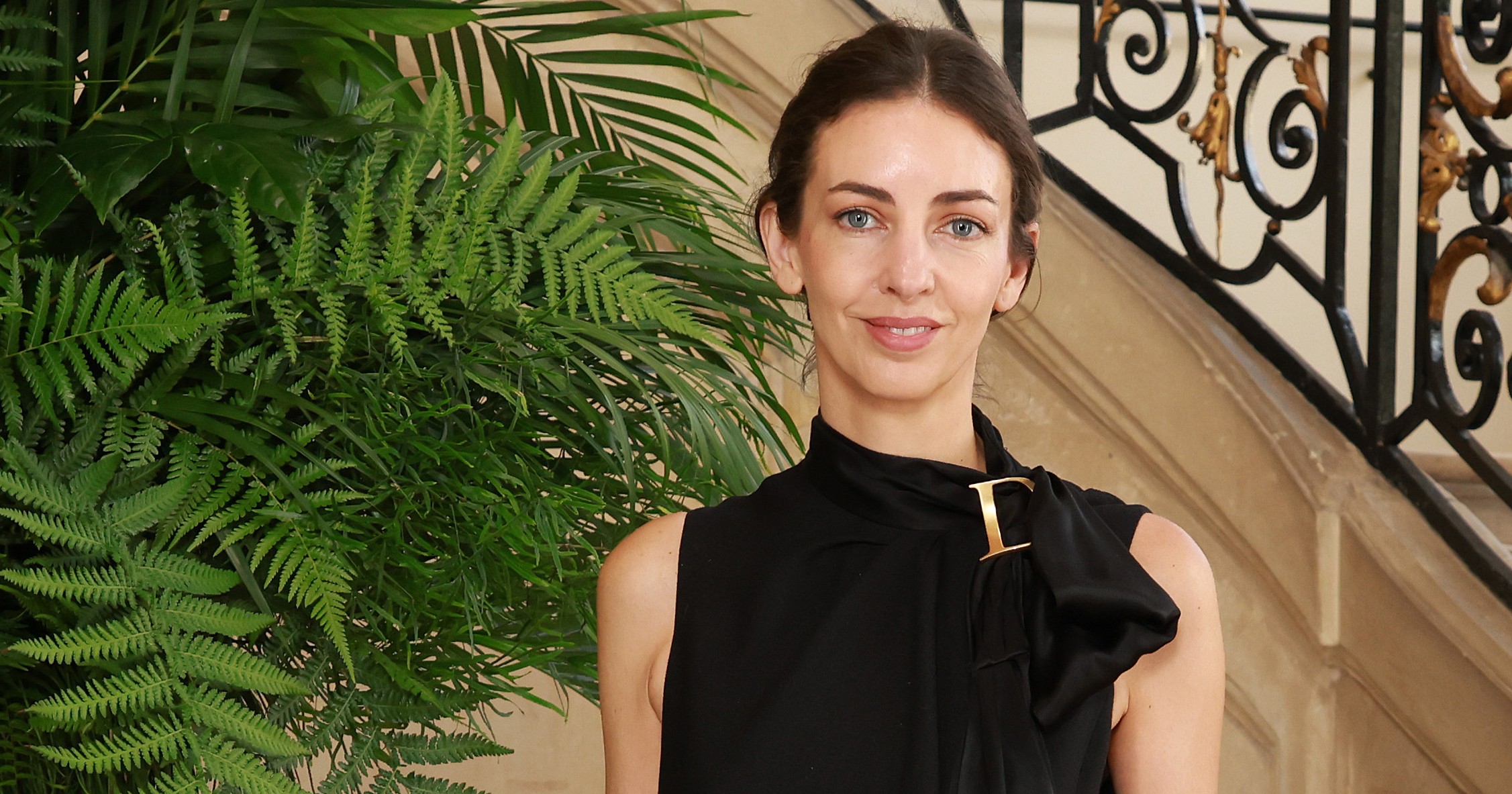 Rose Hanbury in a black sleeveless mini dress with a long satin tie and gold brooch, standing before an ornate wrought-iron staircase in Paris.