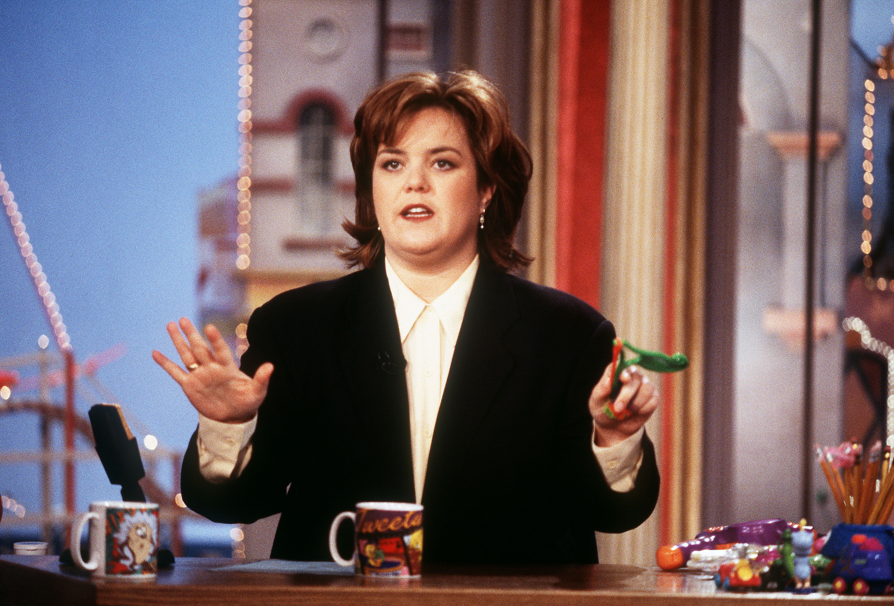 Rosie O'Donnell seated at a talk show desk, gesturing with both hands while holding a small green toy, with mugs and props in front of her.