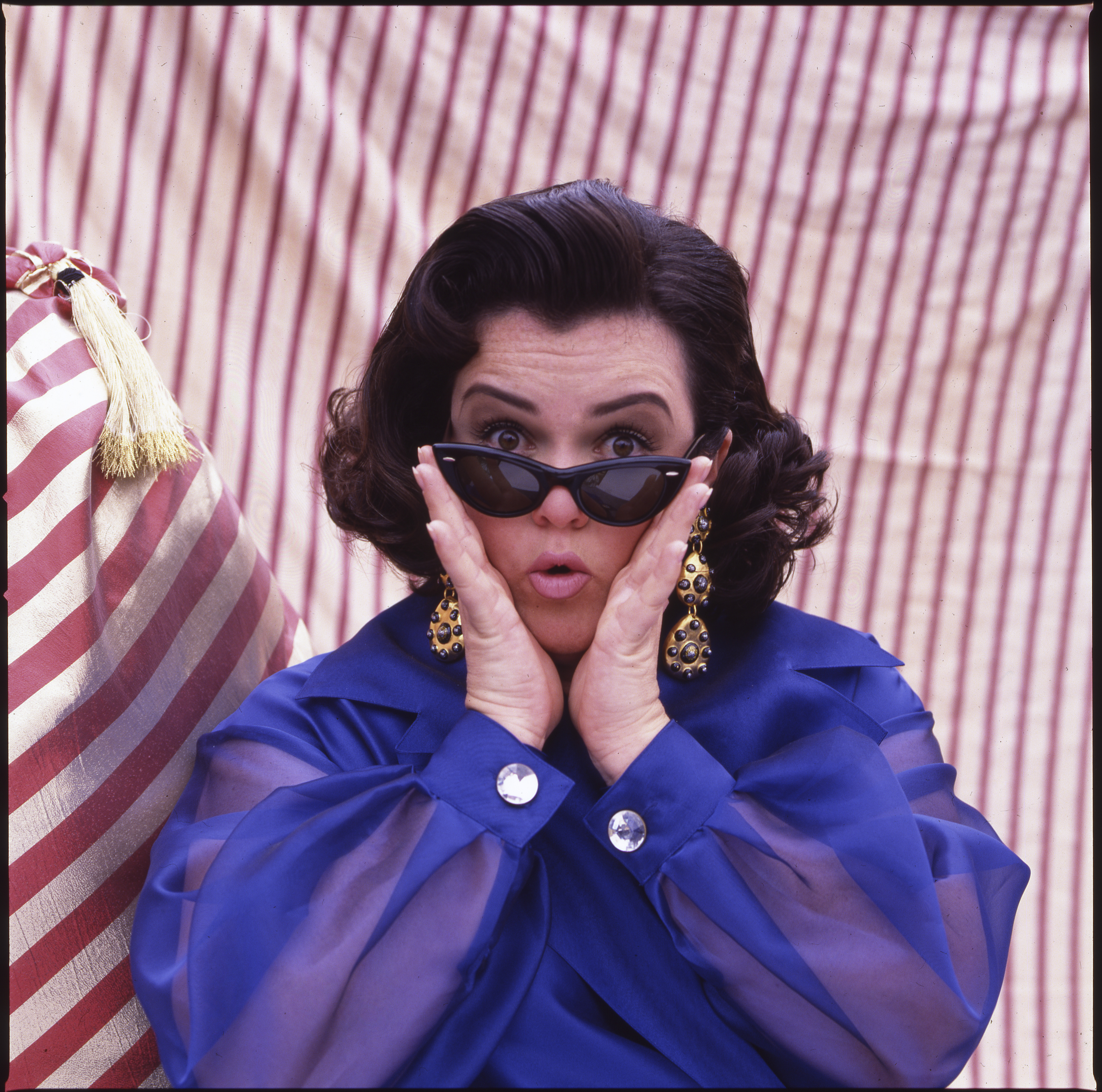 Woman in a blue sheer-sleeve blouse and dark cat-eye sunglasses holds her hands to her cheeks with a surprised expression against a red-and-white striped backdrop.