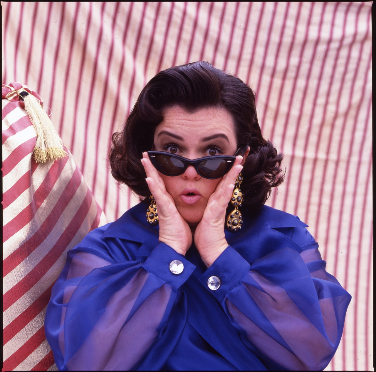 Rosie O'Donnell in a blue blouse and cat-eye sunglasses, hands on cheeks, making a surprised expression against a red-and-white striped backdrop.