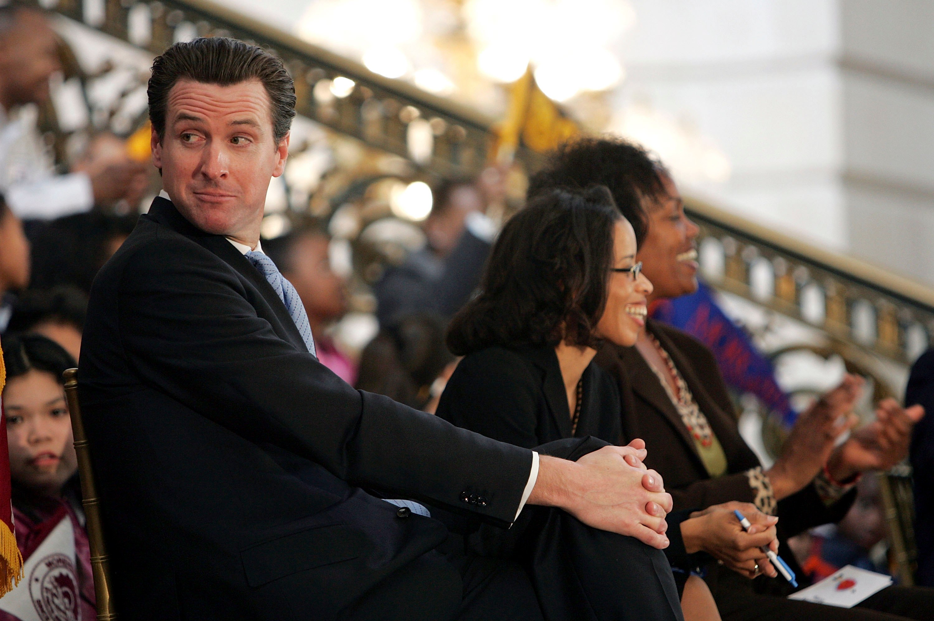 Man in dark suit and blue tie seated in a crowd, turning to look over his shoulder at a formal indoor event with a grand staircase in the background.
