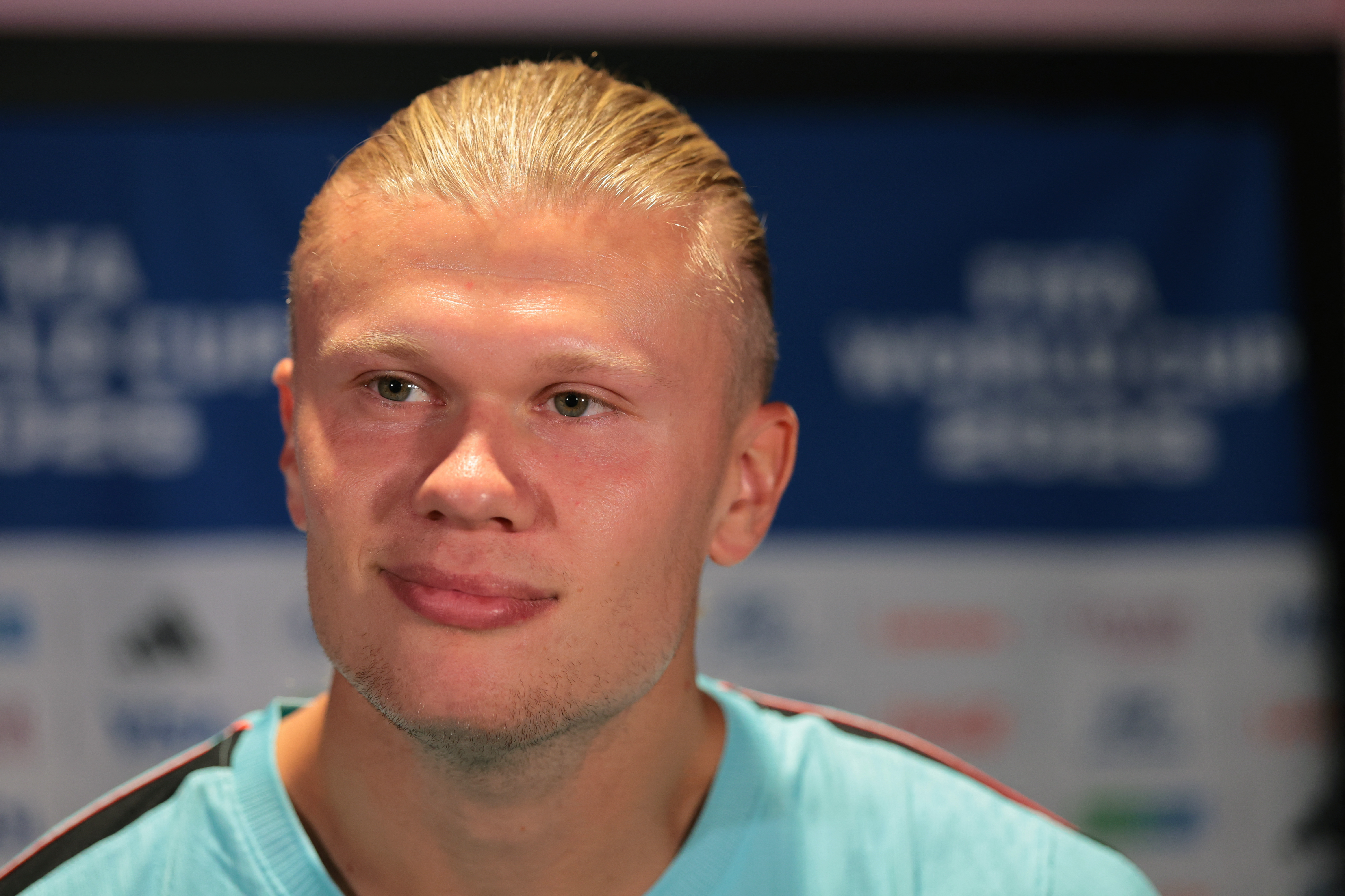 Click to open the image gallery Erling Haaland with slicked-back blond hair in a light blue shirt speaks at a media mixed zone in front of a blue FIFA World Cup backdrop.