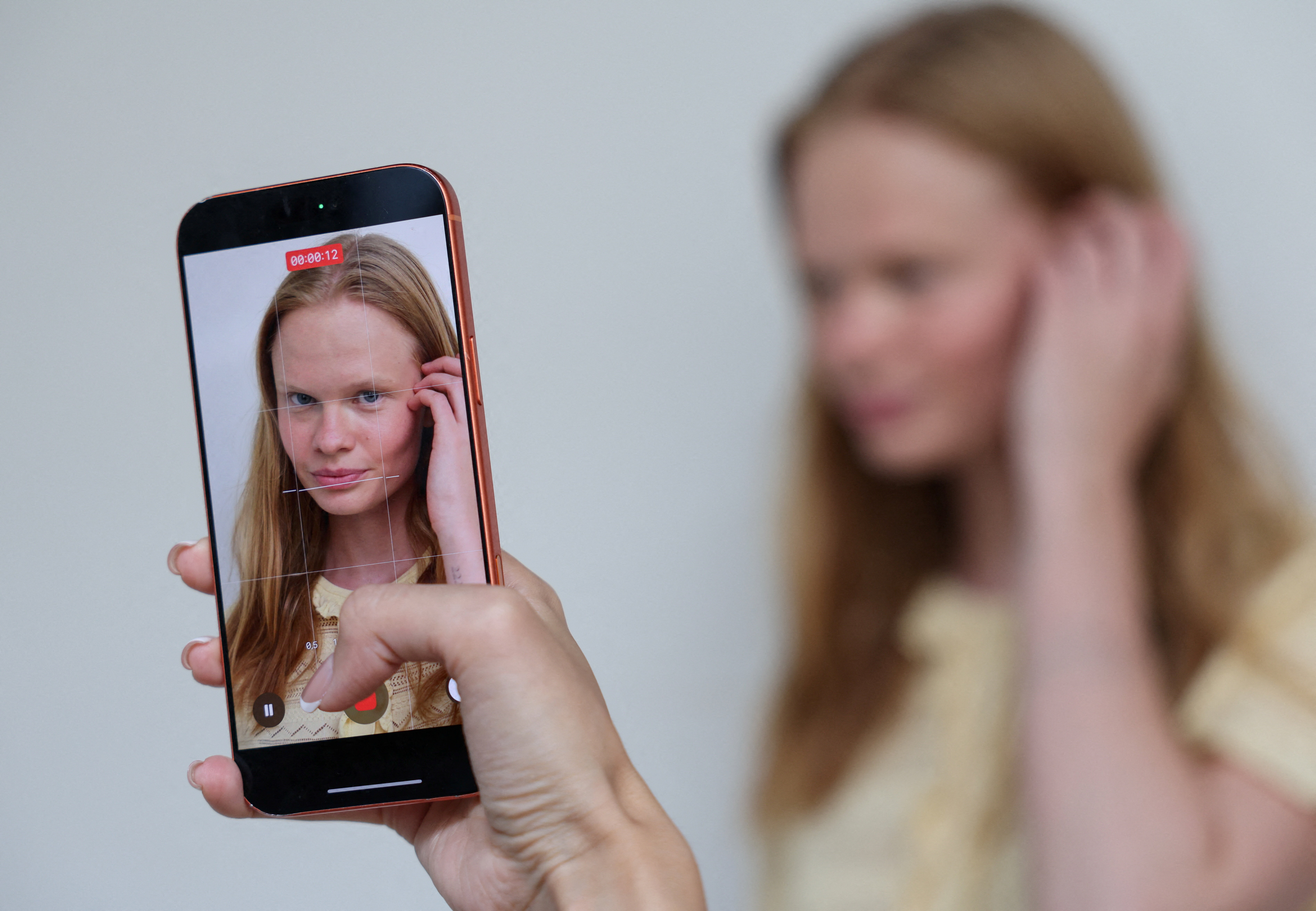 Click to open the image gallery A smartphone held up showing a video recording of a young woman with long blonde hair touching her ear, with the blurred subject visible in the background.