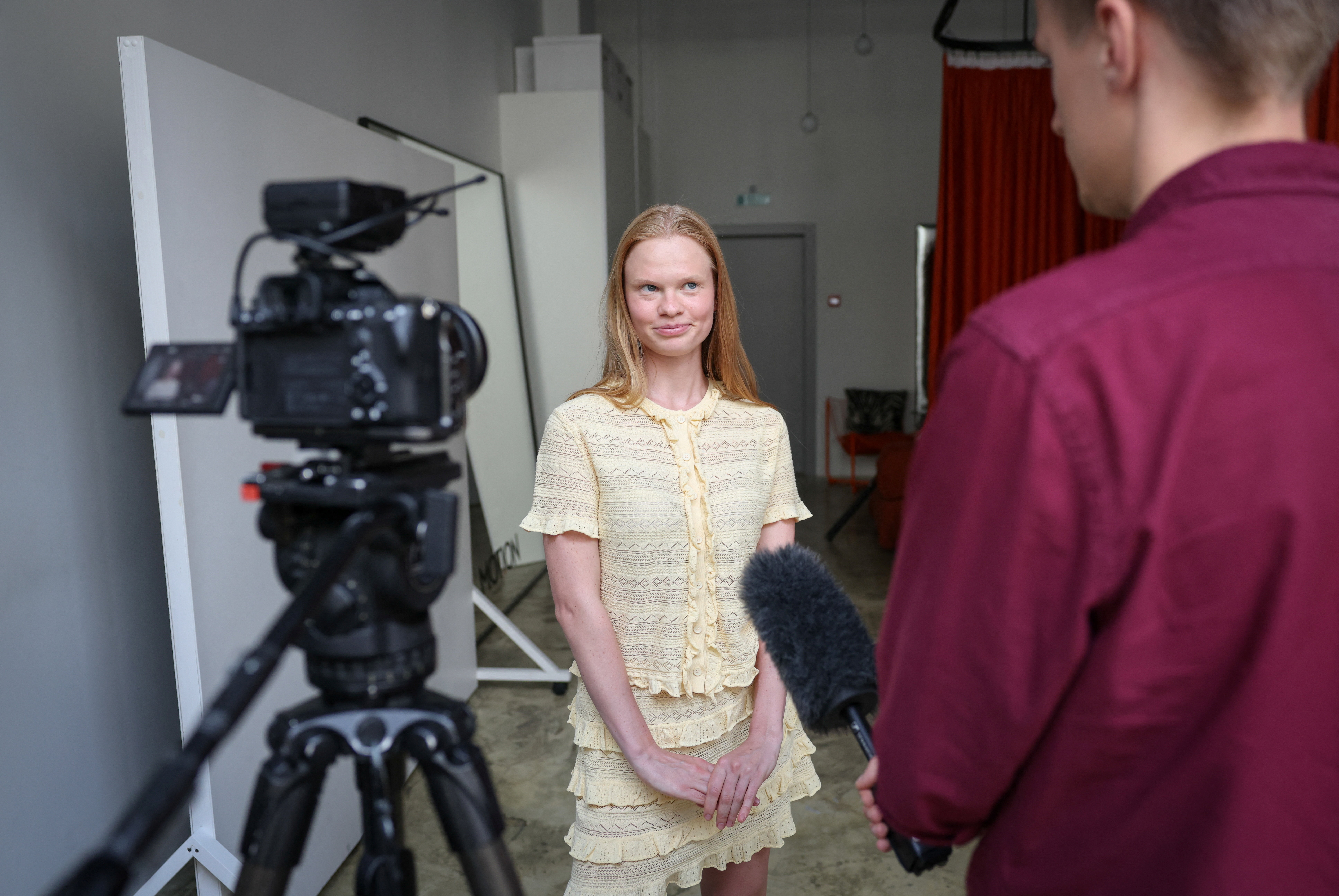 Click to open the image gallery A young blonde woman in a yellow lace outfit stands in a studio being interviewed, with a video camera on a tripod to her left and a person holding a microphone to her right.