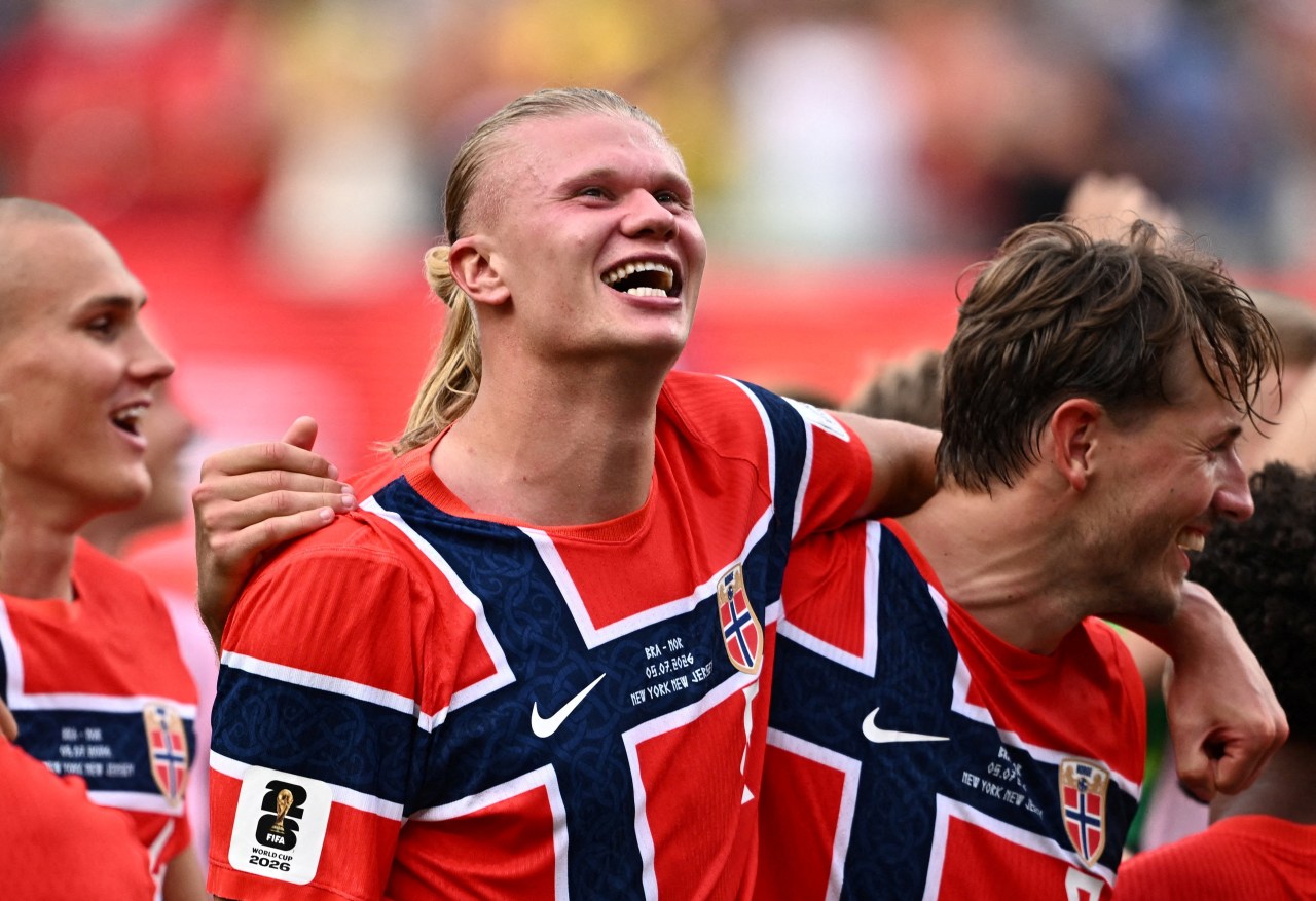 Erling Haaland in a red and navy Norway kit laughs joyfully with teammates on the pitch after a World Cup match.