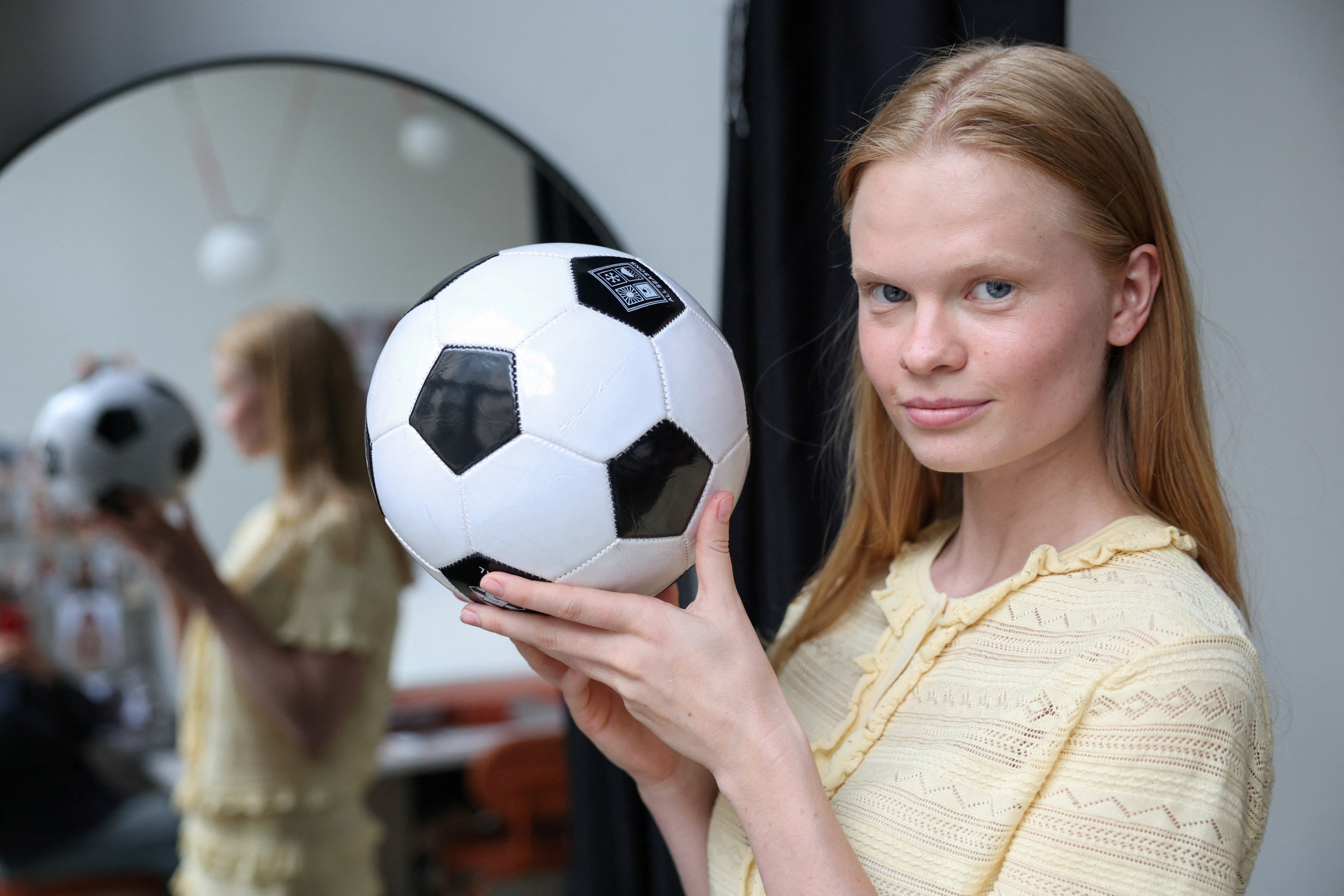 Young woman with long auburn hair holding a black-and-white soccer ball up near her face, her reflection visible in a round mirror behind her.