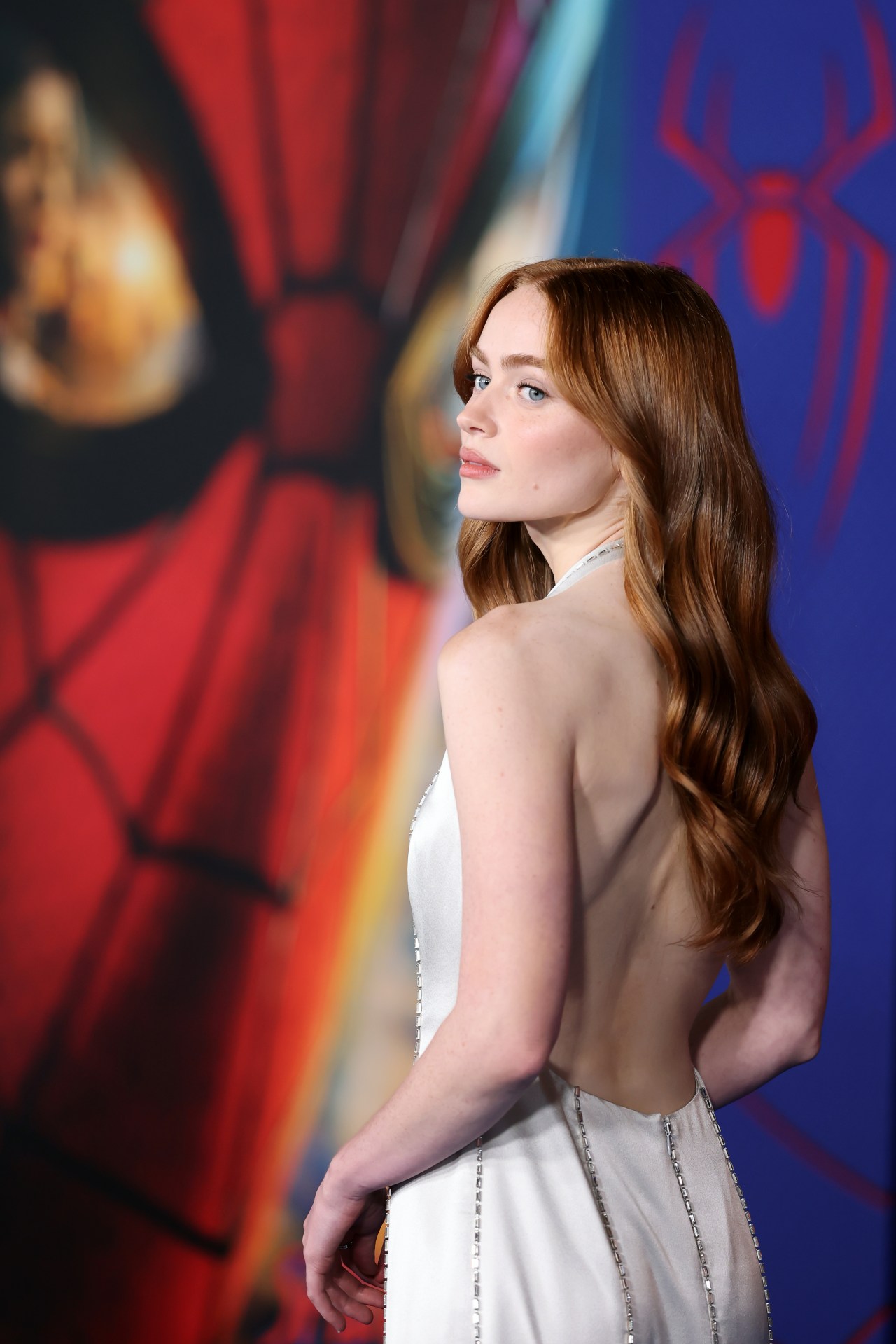Young woman with auburn hair looks back over her shoulder, showing the backless design of her silver satin gown in front of a colorful Spider-Man movie mural.