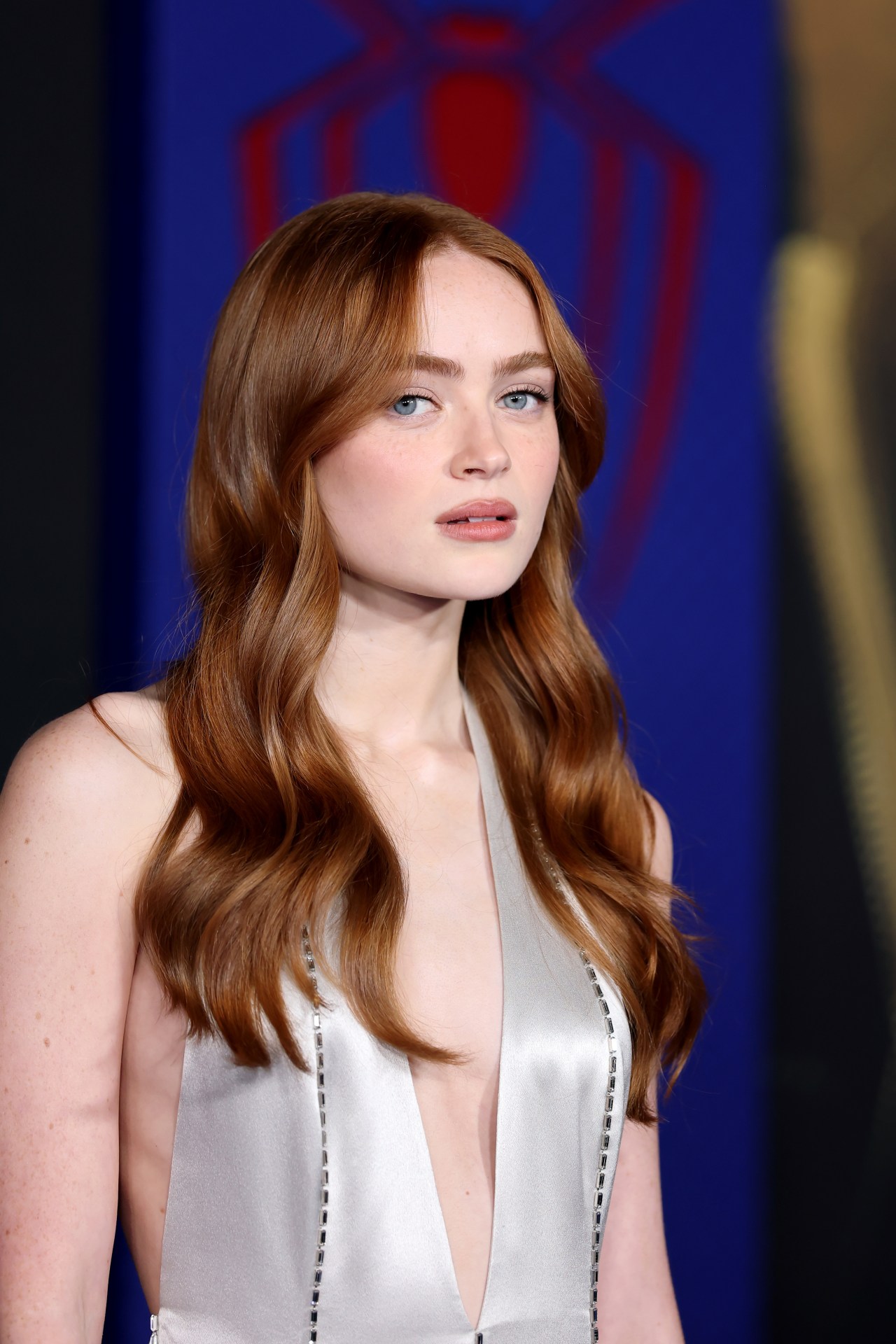 Close-up of Sadie Sink with long auburn wavy hair wearing a deep-V silver satin gown at the Spider-Man: Brand New Day premiere.