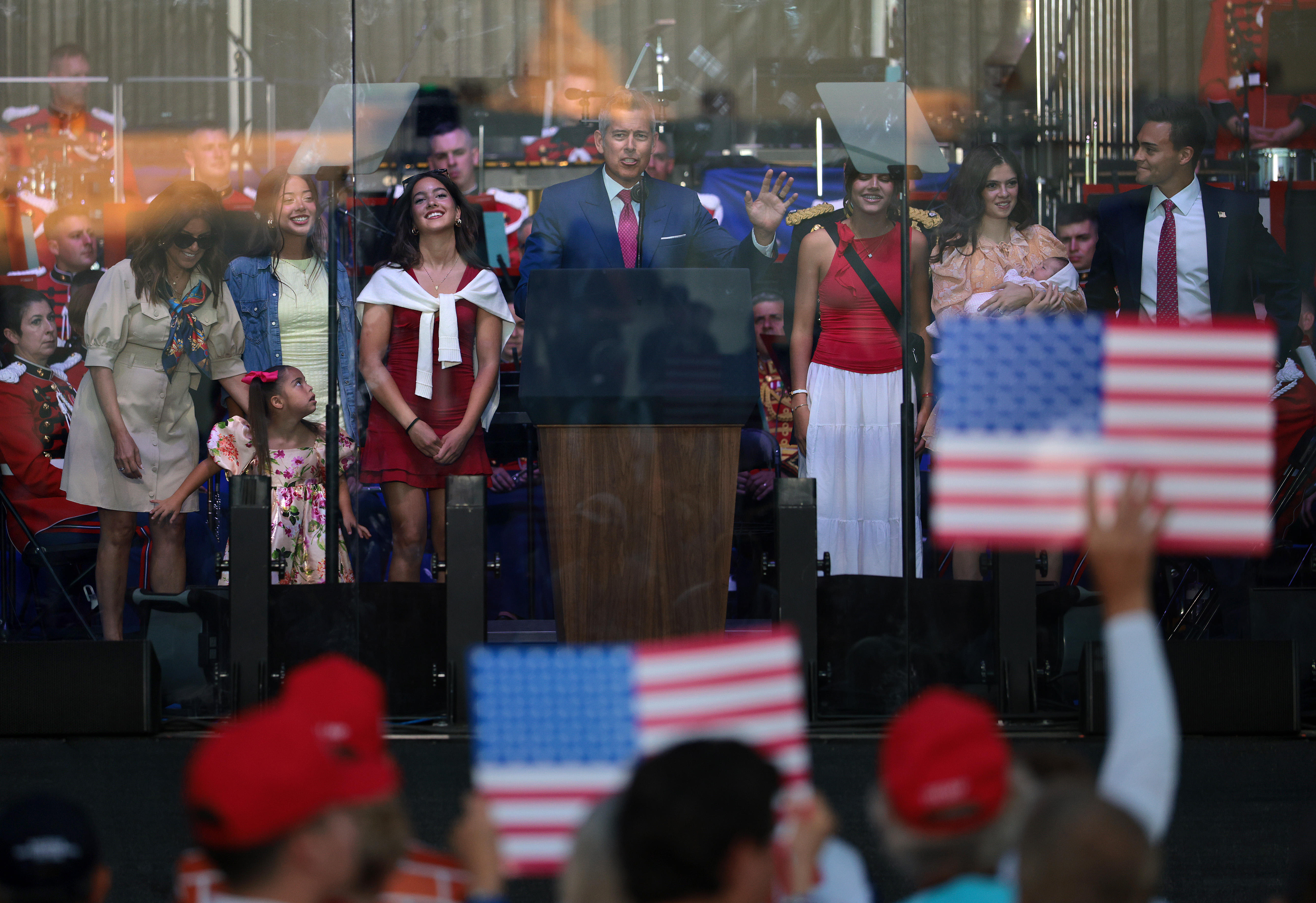 Sean Duffy speaks at a podium on stage surrounded by family members, with audience members waving American flags in the foreground.