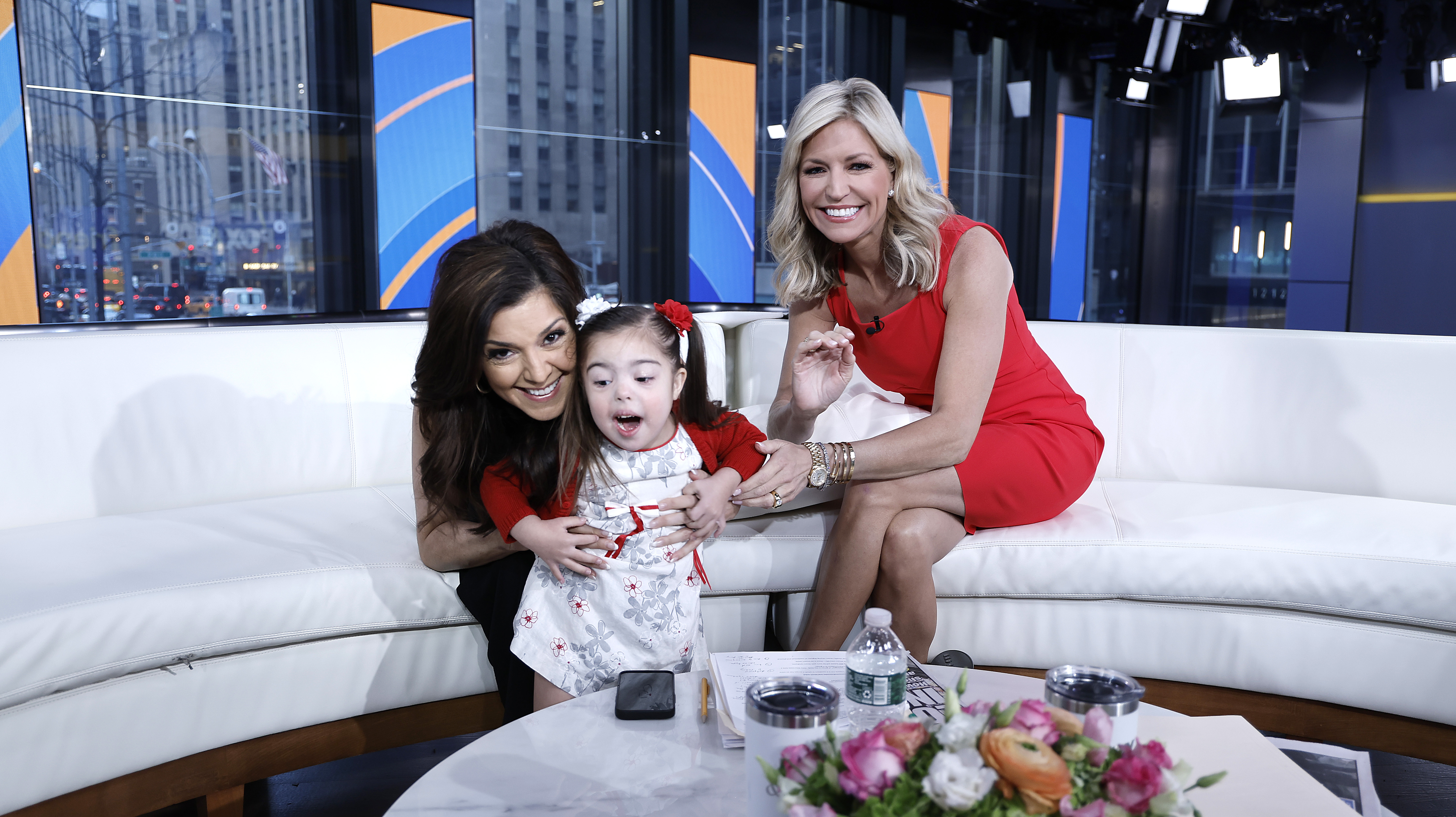 Two women smile on a white studio couch with a young girl with Down syndrome between them on a TV set.