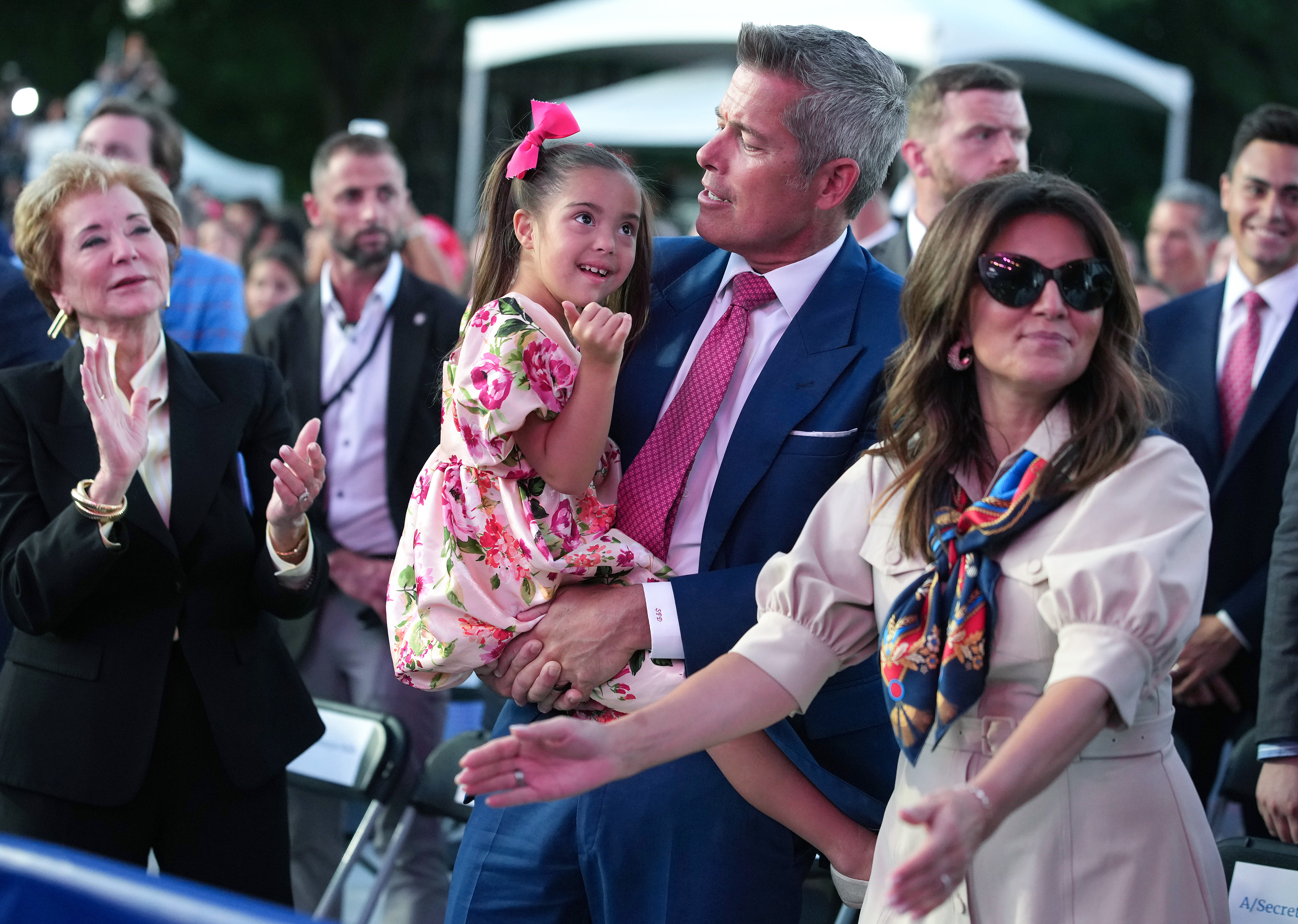 A man in a blue suit holds a young girl in a floral pink dress with a pink bow in her hair at an outdoor rally.