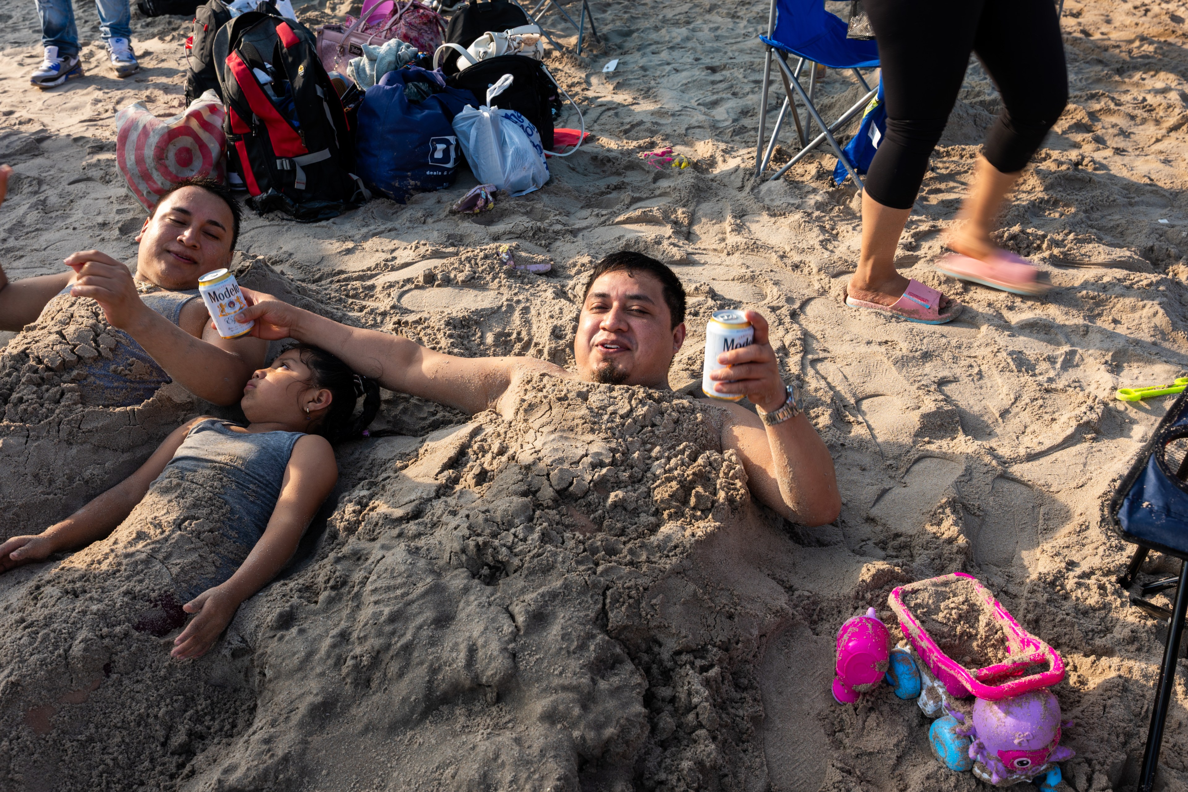 A man buried in sand at a beach holds a can of Modelo beer while a woman and child lie in the sand beside him, surrounded by beach gear.