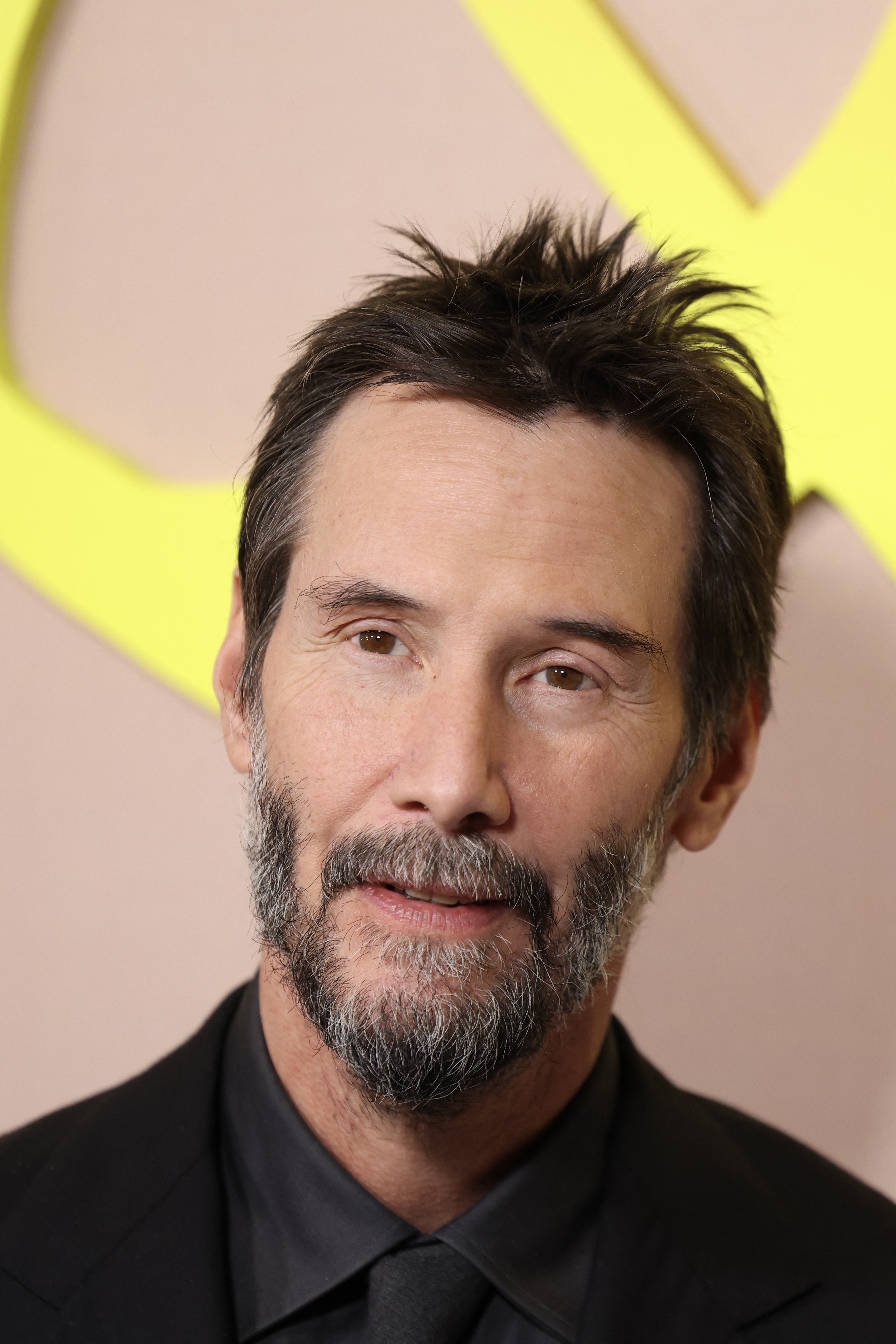 Click to open the image gallery Keanu Reeves with dark spiky hair and a short beard wearing an all-black suit at a premiere event with a yellow logo backdrop.